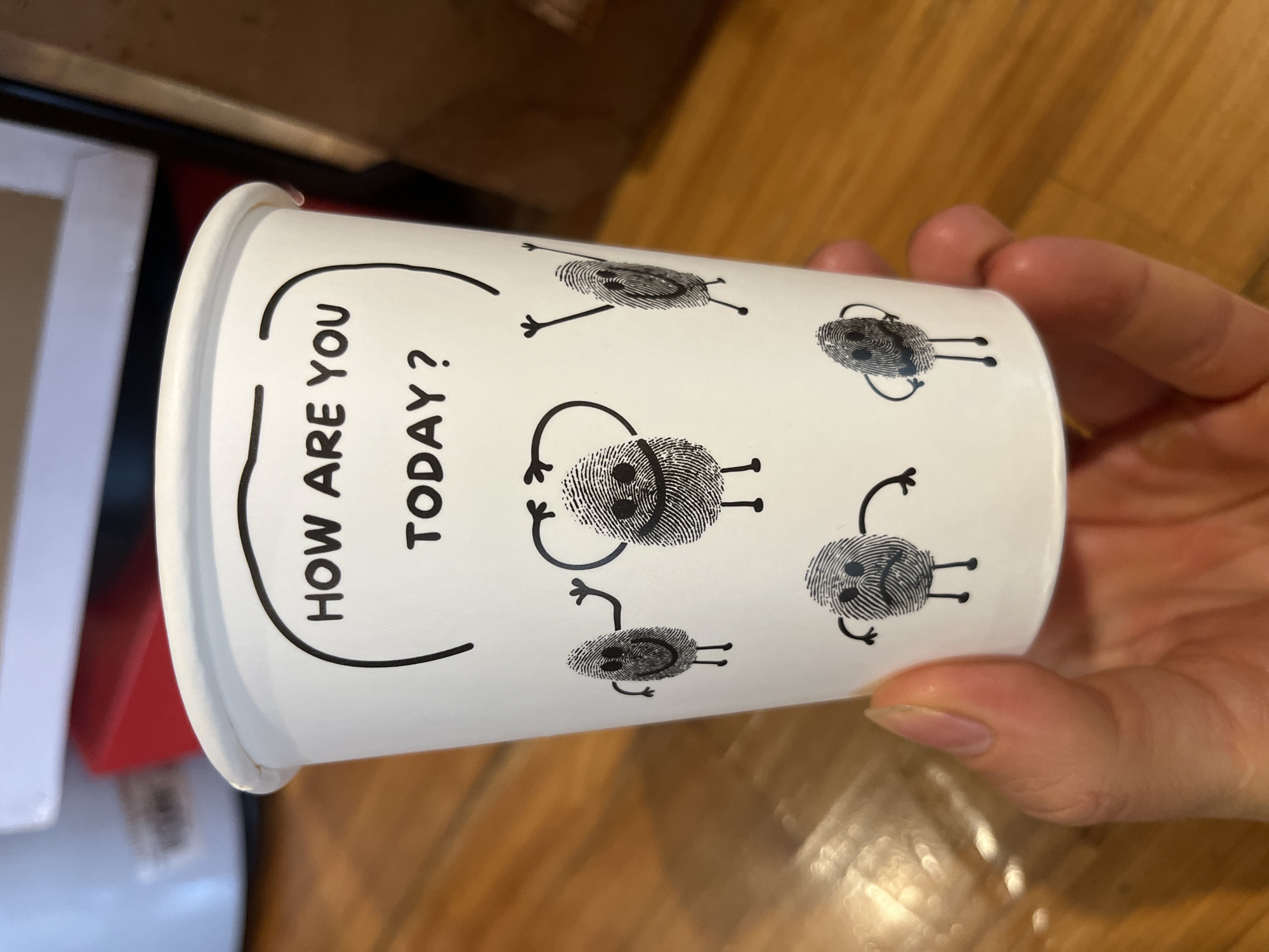 A close-up, eye-level shot shows a person's hand holding a white paper coffee cup. The cup has a black line drawing of a fingerprint with arms and legs on it, and the words "HOW ARE YOU TODAY?" are written in black text above the fingerprint character. The fingerprint characters are in various poses and facial expressions, depicting different emotions. The background is blurred and appears to be an indoor setting with wooden flooring and some indistinct objects. The specific location in Phu Thuy Ward, Vietnam, is not discernible from the image.