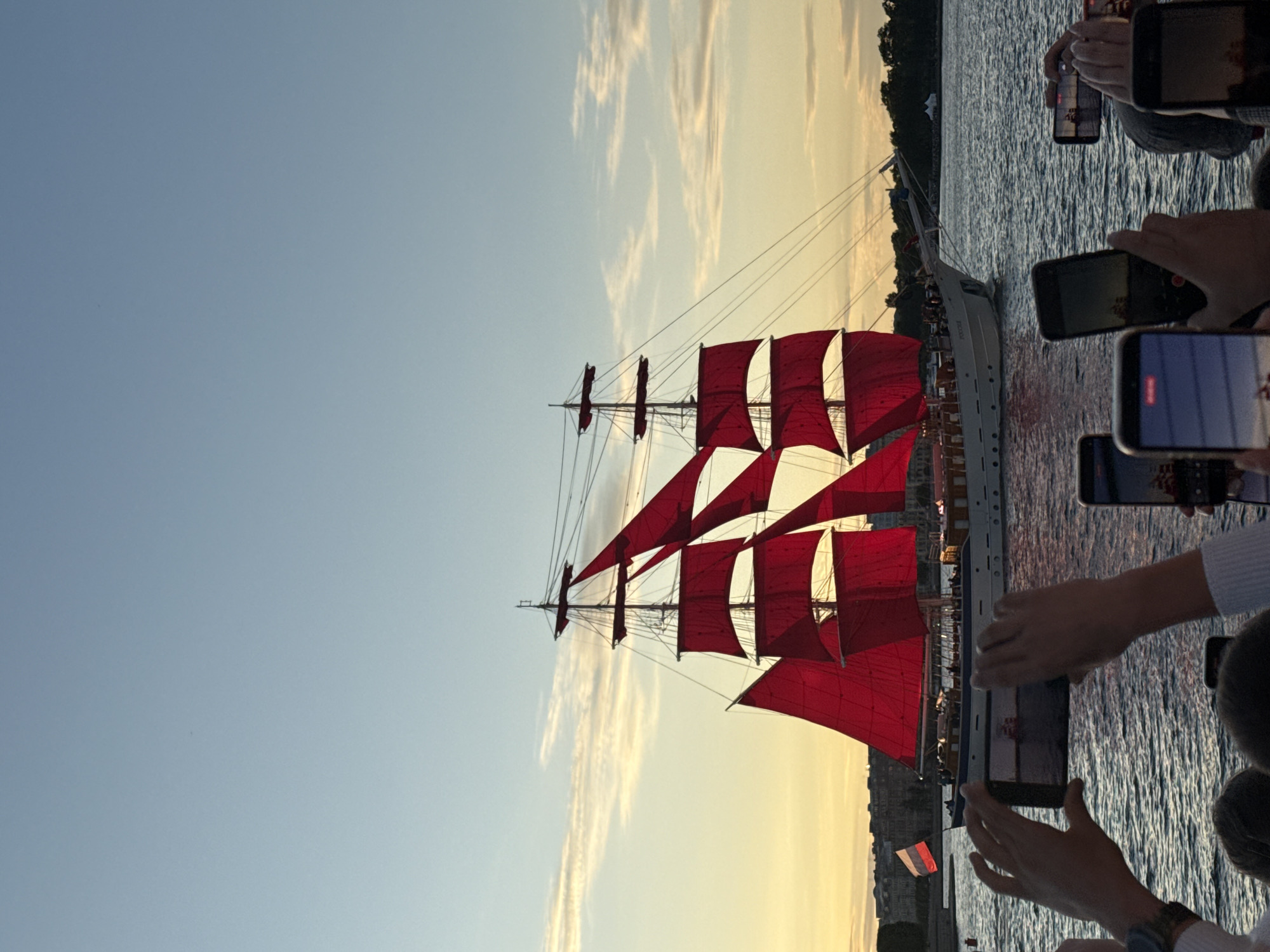 This image depicts a historic sailing ship with vibrant red sails navigating through a body of water, likely a river or canal, in Saint Petersburg, Russia. The ship, a prominent feature, is a tall ship with multiple masts and rigging, its sails catching the soft light of either sunrise or sunset, creating a dramatic visual. In the foreground, a group of individuals are gathered, their hands raised to capture the scene with their mobile phones, indicating a public event or spectacle. The background shows a city skyline with buildings and trees, consistent with the architecture of Saint Petersburg, and a clear sky with subtle cloud formations. The presence of a Russian flag on a smaller vessel near the main ship further confirms the location. The overall atmosphere is one of awe and observation, with the ship serving as the focal point of the captured moments.