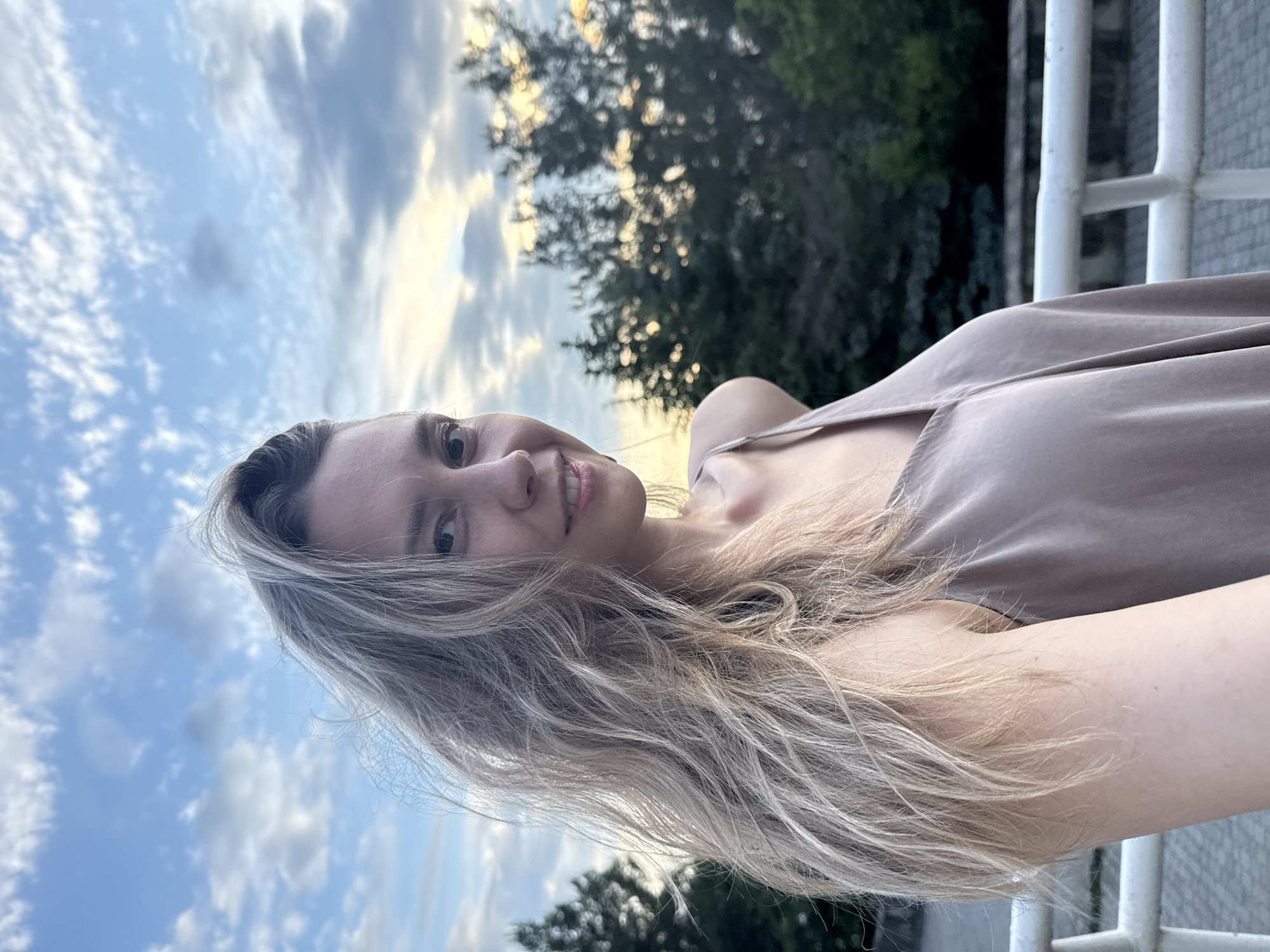 A young woman with long blonde hair is captured in a selfie outdoors, with a cloudy sky and trees in the background. She is wearing a light-colored, spaghetti-strap dress. The setting appears to be a bridge or elevated walkway, indicated by a white railing in the foreground. The woman is looking towards the camera with a slight smile. The location is identified as the city of Nebug, Russia.
