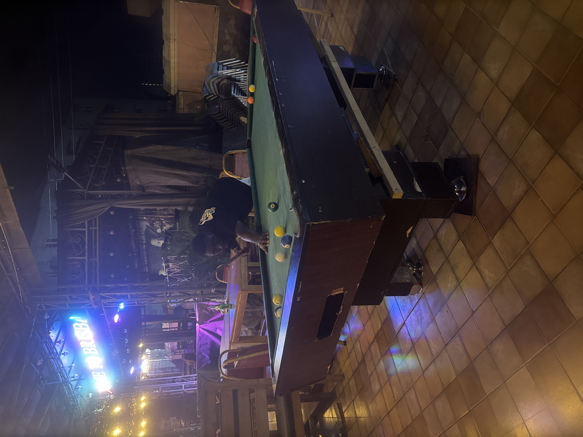 A man is playing pool at night in Nkpor, Nigeria. The scene is outdoors, likely a bar or a recreation area, with a neon sign in the background that reads "WELCOME TO BRISBANA". The pool table is green with several balls on it. The man is wearing a black shirt with a graphic design and is in the process of hitting a ball with his cue. The ground is tiled and reflects the colored lights from above. There are tables and chairs visible in the background, along with stacked chairs and some draped fabric. The overall atmosphere is casual and social.