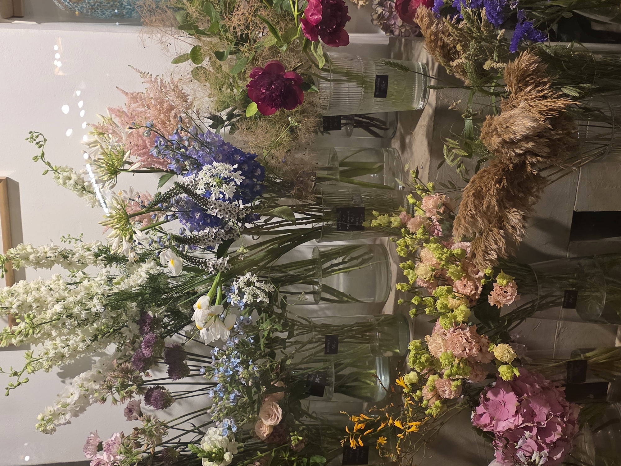 The image shows a display of various flowers in glass vases. The flowers are arranged in a variety of colors and types, including white delphiniums, pink hydrangeas, purple alliums, and yellow spray roses. The flowers are in different stages of bloom, and some are in bud. The display is set against a plain white background, with some hints of a wooden beam at the top. The lighting is soft and diffused, creating a calm and serene atmosphere. There are price tags visible on some of the vases, indicating that this is likely a flower shop or market.