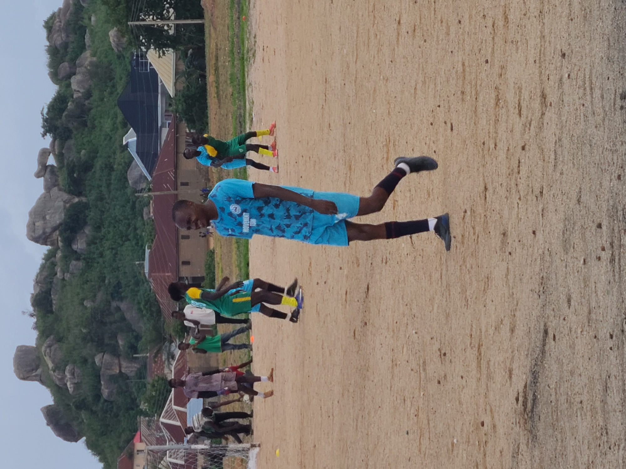 The image depicts a group of young men on a sandy field, likely a sports ground, in Jos, Nigeria. The scene suggests a training session or a casual game of football, as several individuals are dressed in sportswear. One person in the foreground is in motion, mid-stride, wearing a light blue jersey with a camouflage pattern and matching shorts, along with black socks and dark shoes. Other individuals in the background are also engaged in activity, some in green and yellow jerseys.
The setting is rural, with a prominent rocky hill covered in lush green vegetation dominating the background. Several buildings with corrugated iron roofs are visible amongst the trees, indicating a nearby settlement. The ground is covered in light-colored sand with scattered small stones. The lighting suggests it is daytime, possibly late afternoon, given the soft shadows and the overall illumination. The atmosphere appears to be casual and energetic, typical of a sporting event. No discernible text is visible in the image.