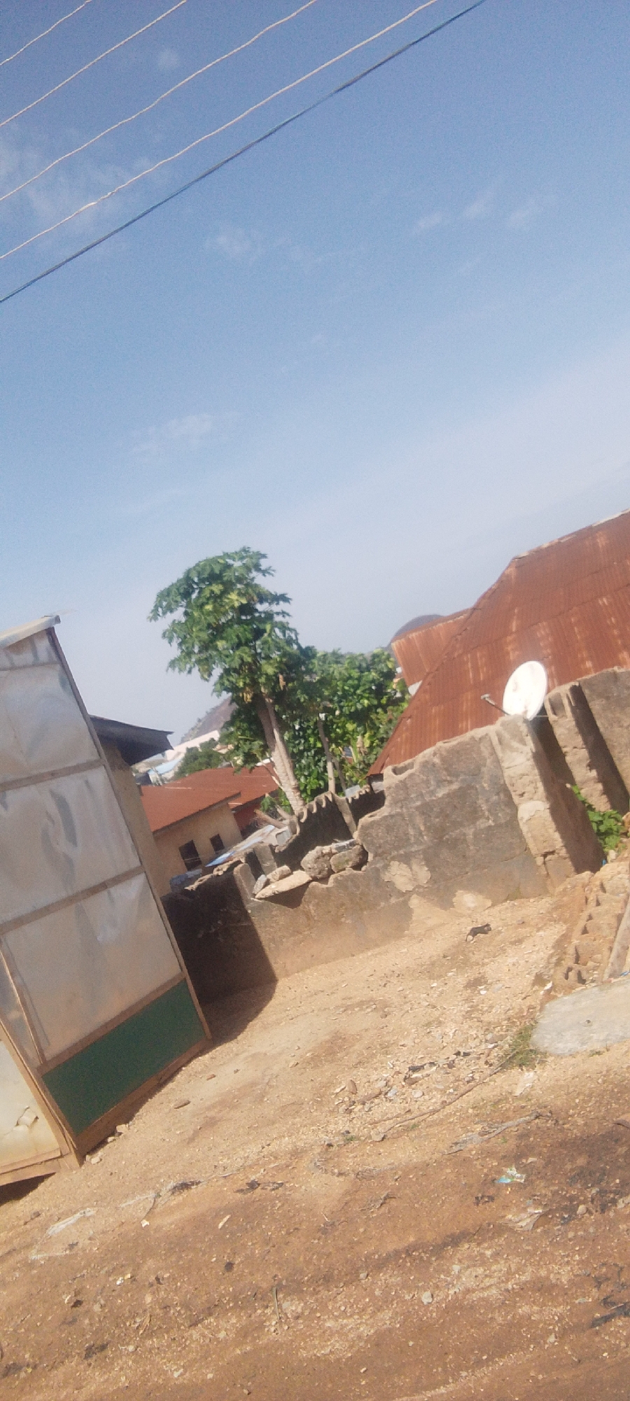 The image shows a scene in Madalla, Nigeria, characterized by a dusty, uneven ground with scattered debris. In the foreground, a large, partially constructed structure with metal sheeting and a green lower section is visible on the left. Behind it, a concrete wall, partially collapsed and containing rubble, forms a boundary. A satellite dish is mounted on top of this wall, facing left. Beyond the wall, a rust-colored corrugated metal roof of a building is prominent. A tree with lush green foliage stands between the structures. In the background, several buildings with reddish-brown roofs are clustered together, and a distant hilly landscape is visible under a clear blue sky with thin, horizontal power lines crossing the upper portion of the frame. The overall impression is of a developing or informal settlement, with evidence of ongoing construction and some disrepair.