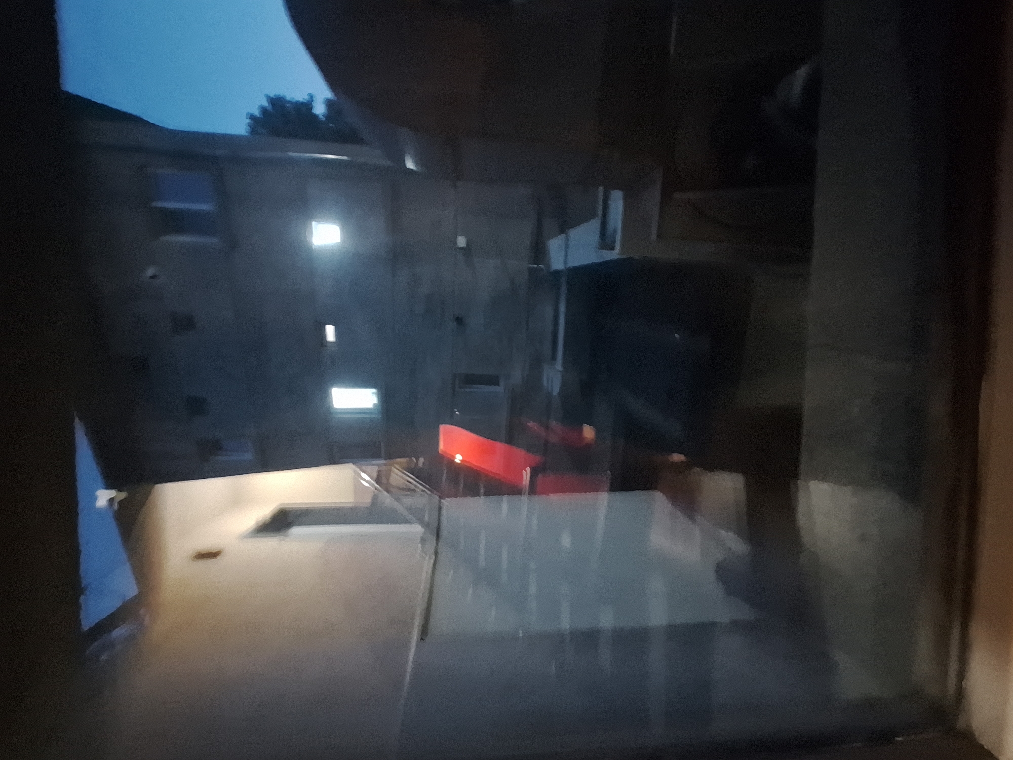 The image depicts an outdoor scene at dusk or night, likely in Varna, Bulgaria. In the foreground, a window pane reflects the scene with a bluish tint and some glare. Beyond the window, a building with illuminated windows is visible. To the left, a lighter-colored building with a window and a dark opening is present. A red, angular object, possibly a slide or a part of a structure, is positioned between the buildings. Stairs are also visible leading up to the lighter building. The overall environment appears to be an urban or suburban setting. The scene is dimly lit, suggesting it is after sunset. There are no discernible individuals or specific actions taking place.