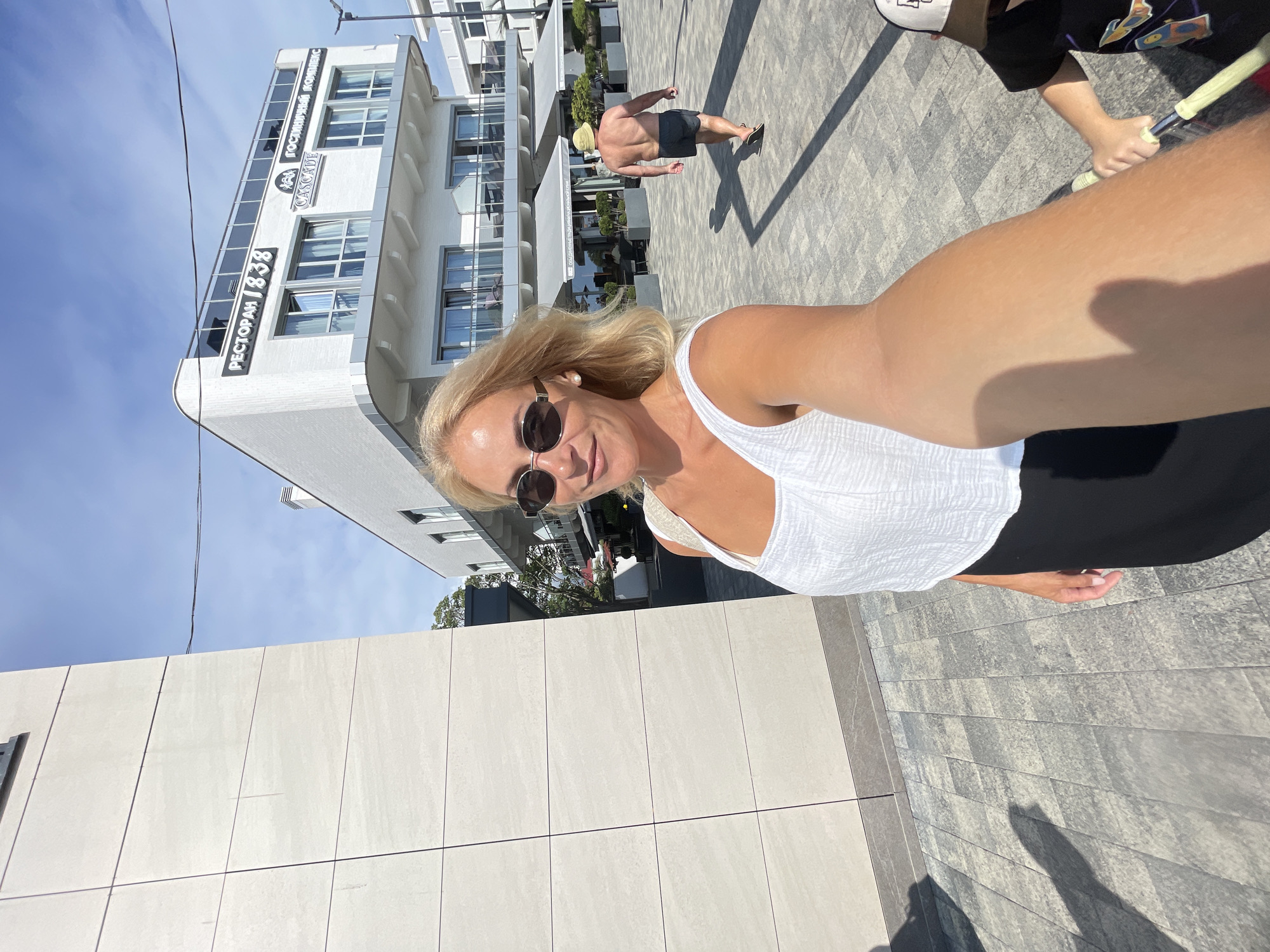 This is a candid, selfie-style photograph taken outdoors on a sunny day. The scene appears to be a modern urban area, possibly a resort or promenade, with a contemporary building in the background and a paved walkway.
The primary subject is a woman with blonde hair, wearing sunglasses and a white top. She is smiling and looking towards the camera, suggesting a cheerful or relaxed mood. Her arm and shoulder are prominently featured in the foreground due to the selfie perspective.
In the middle ground, a man is visible walking away from the camera. He is wearing a straw hat and dark shorts, and his back is to the viewer, showcasing his tanned physique. Another person, partially visible, is holding a yellow object, possibly a racket, further in the background.
The building in the background is white with multiple stories and large windows. Visible text on the building includes "РЕСТОРАН 1838" (RESTAURANT 1838) and "ГОСТИНИЧНЫЙ КОМПЛЕКС CASCADE" (HOTEL COMPLEX CASCADE). The presence of "Sochi, Russia" as location context indicates this is likely a location along the Black Sea coast.
The lighting suggests it is daytime, with distinct shadows cast on the ground, indicating the sun is relatively high in the sky. The overall atmosphere is bright and summery.