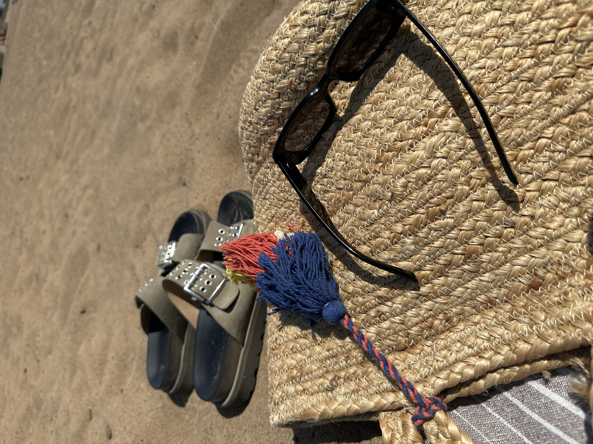 The image shows a beach scene, likely taken during the day. The foreground features a pair of light brown sandals with silver buckles resting on the sand. Next to the sandals is a woven straw bag. On top of the bag are black sunglasses and a decorative tassel with blue and red strands. The environment consists of sand, indicating a beach or sandy area. There is no indication of this image being taken in the city of Chaikovsky, Russia. No people are visible in the image.