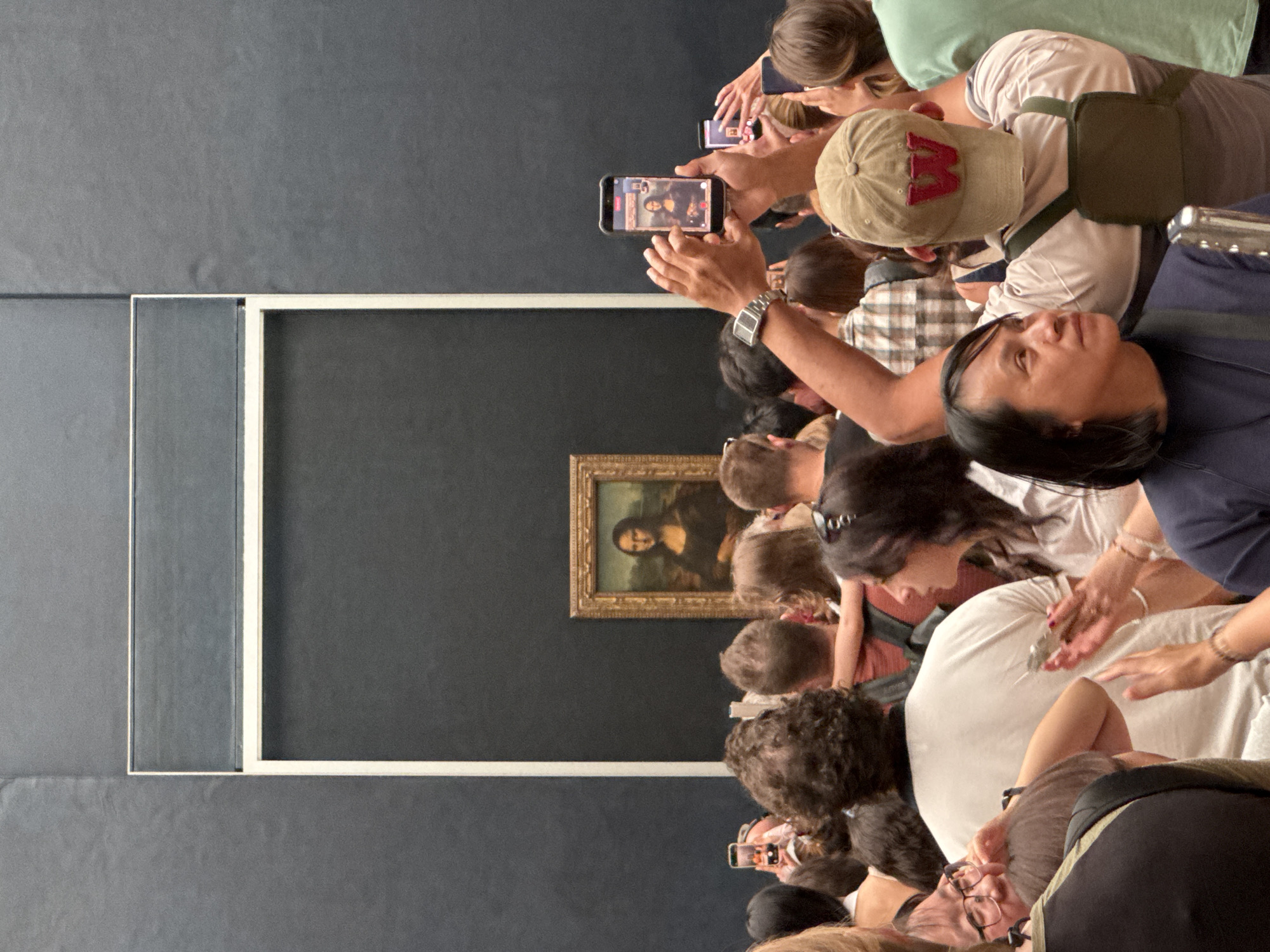 This image depicts a crowd of people gathered in front of the Mona Lisa painting at the Louvre Museum in Paris, France. The painting is housed in a gold frame and displayed against a dark background. Many individuals in the foreground are holding up smartphones to take pictures or record videos of the artwork. The crowd appears dense, with people of various ages and backgrounds. The atmosphere seems to be one of excited tourism, with a focus on capturing the iconic painting through personal devices. The lighting suggests an indoor setting, consistent with a museum environment.