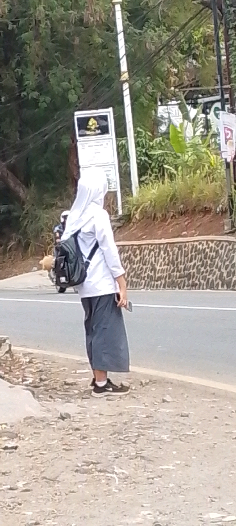 In the city of Cilaku, Indonesia, a person wearing a white hijab and a white shirt with a gray skirt stands on the side of a road. They are carrying a dark backpack and holding a mobile phone. A motorcycle is visible behind them, and the background shows trees, a sign, and a stone wall. The ground is covered in dirt and debris.