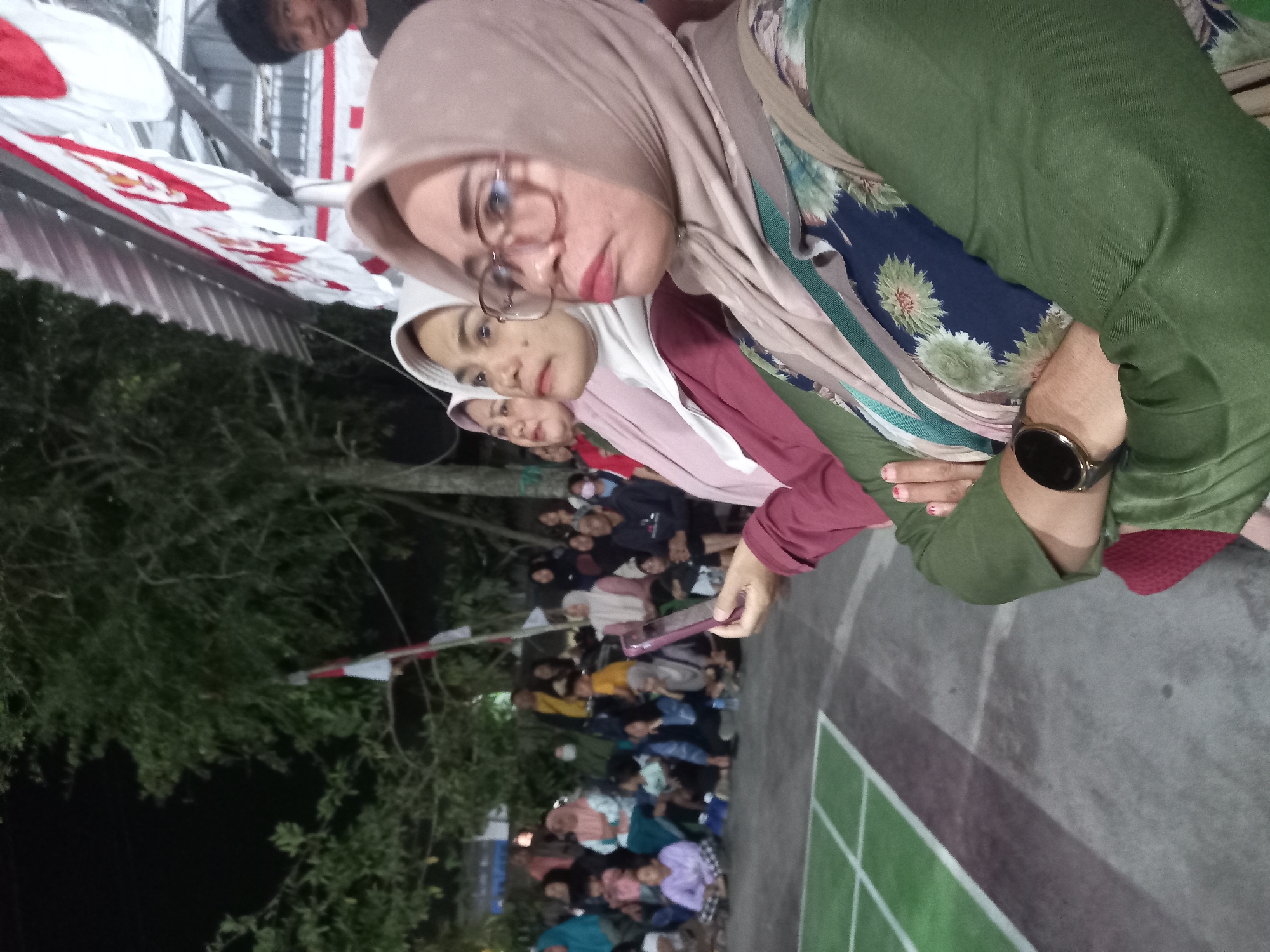 In Banyumulek, Indonesia, at night, a group of people are gathered outdoors. In the foreground, a woman wearing glasses and a light brown hijab is looking to the right, with her arms crossed. She is wearing a green long-sleeved shirt with a floral pattern underneath. To her left, two other women in hijabs are visible, one in a pink hijab and the other in a white hijab, both looking in the same direction as the first woman. Behind them, a crowd of people, including children and adults, are seated and standing. Some are watching an event or performance, while others are looking around. A red and white Indonesian flag is visible hanging in the background, along with some trees and lights. The ground is a paved surface with green lines marking a court, possibly for a sport. The atmosphere appears to be festive, possibly a local event or celebration.