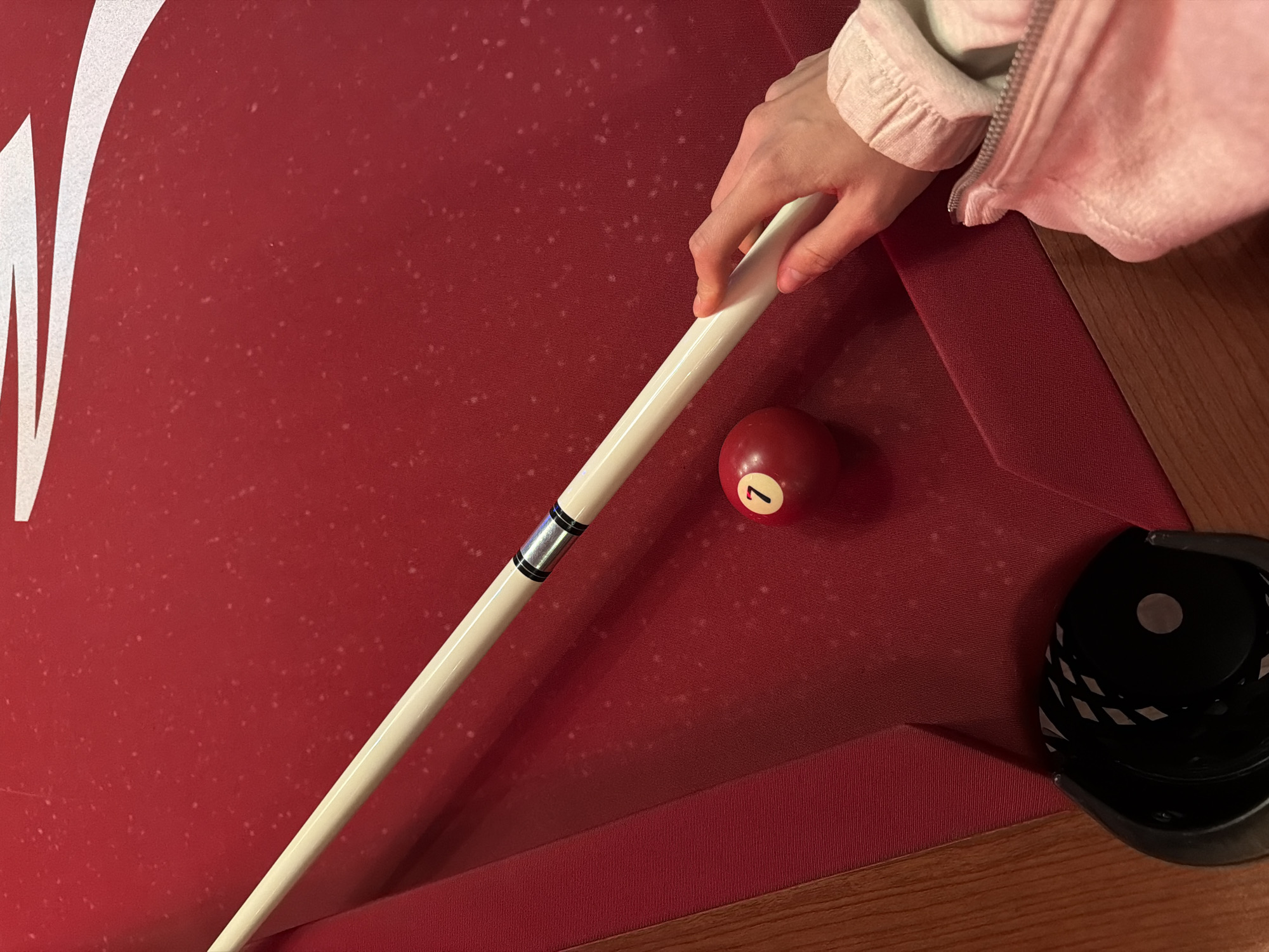 This close-up image captures a person's hand preparing to play billiards on a red felt pool table. The main subject is a hand, with light skin tone, holding a cream-colored pool cue. The hand is positioned over the cue, as if steadying it for a shot. The person is wearing a light pink, long-sleeved garment with a ruffled or elasticized cuff and a silver zipper visible further up the arm.
On the red felt of the pool table, a single red billiard ball with the number "7" in a white circle is visible. The cue is aligned towards this ball. The red felt surface is speckled with numerous small, light-colored dots. In the upper left corner, part of a large white letter or decorative symbol, resembling a "W" or "M," is visible on the felt. The edge of the pool table, made of dark wood, is seen at the bottom right. Next to the wooden edge, a black object with white markings, possibly a pool chalk holder or part of a cue rack, rests on the wood. The scene depicts an indoor setting, likely a game room, with the activity of playing pool about to commence. The specific location is Krasnoyarsk, Russia, though no visual cues within the image confirm this.