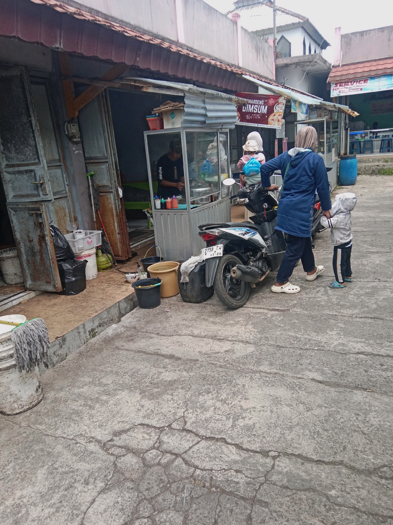 A mother, wearing a blue hoodie and a hijab, stands beside a scooter in what appears to be a street-side food stall in Bujungsari, Indonesia. She is holding the hand of a young child, who is wearing a grey hooded jacket and black pants. The child is facing away from the camera, looking towards the food stall. The stall, labeled "WARDENG DIMSUM BATUR," is an open-air establishment with a metal roof and counter. A person, likely the vendor, is visible inside the stall. Several buckets, a gas cylinder, and cleaning supplies are scattered on the ground near the entrance of an adjacent building, which has large metal doors. A sign above the stall reads "SERVICE HPP BISA DITUNGGU" with smaller text below. The ground is paved with concrete that shows signs of wear and cracking. The overall scene depicts a casual, everyday moment at a local eatery in an Indonesian city.