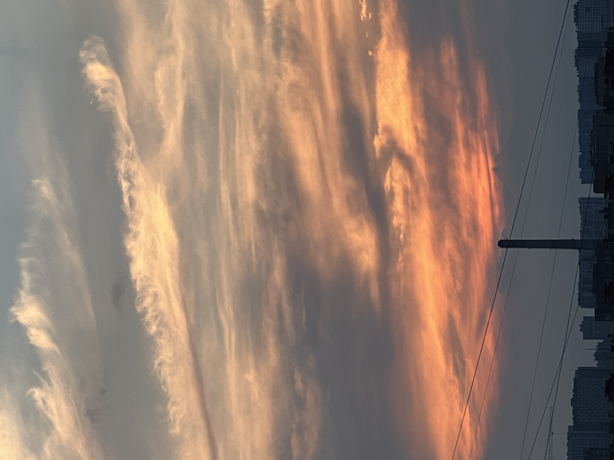 The image captures a dramatic sky over a city skyline, likely in Unknown, Russia, at sunset or sunrise. The sky is filled with swirling clouds painted in hues of orange, pink, and grey, creating a dynamic and textured celestial canvas. Wispy streaks of lighter clouds contrast with darker, more dense formations.
Below the sky, a silhouetted cityscape is visible. Prominent features include a tall, cylindrical industrial chimney, suggesting a factory or power plant. Several buildings of varying heights and architectural styles form the urban landscape. Power lines crisscross the lower portion of the image, adding a layer of man-made infrastructure to the scene. A construction crane is faintly visible on the left side of the frame.
The lighting suggests either the early morning or late evening, with the warm tones of the sun illuminating the clouds from below or behind. The overall impression is one of natural beauty juxtaposed with urban industrial elements. There are no people or visible activity depicted in the image.
