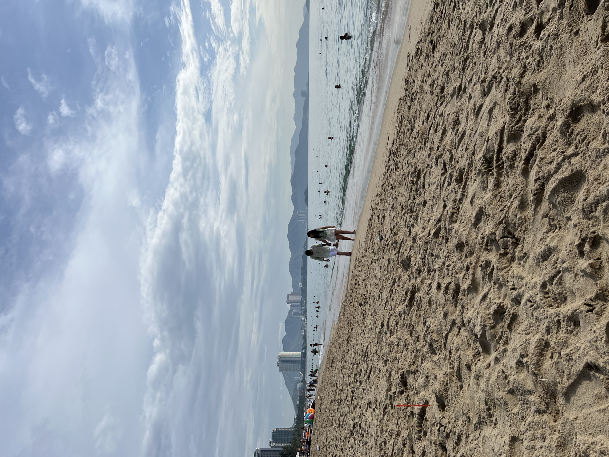 The image depicts a beach scene in Nha Trang, Vietnam, under a partly cloudy sky. In the foreground, the sandy beach shows footprints and textured surfaces. A red marker is visible in the sand on the left side.
Along the shoreline, two individuals, a man and a woman, are walking hand-in-hand. The man is wearing a light-colored shirt and shorts, and the woman is in a light-colored dress. Further along the beach, a group of people are visible, some engaged in activities in the shallow water, while others are closer to the shore. Inflatable rings and other beach accessories are scattered near the waterline.
In the background, the sea extends towards the horizon, with many people swimming and wading in the water. The coastline is lined with tall buildings and mountains, characteristic of the Nha Trang city skyline. The overall atmosphere suggests a relaxed day at the beach.