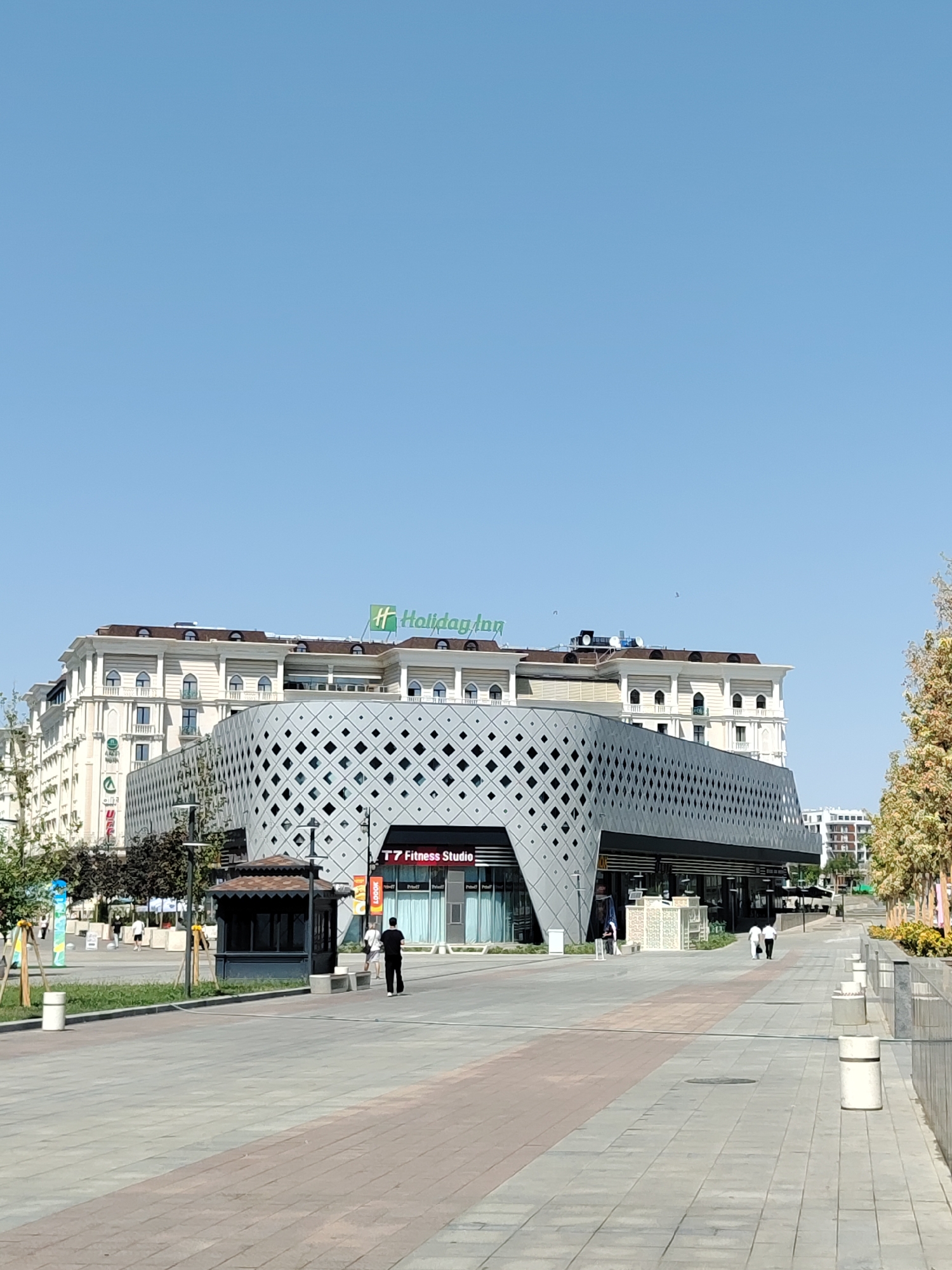 The image shows a modern commercial and hotel complex in Tashkent, Uzbekistan, specifically the Holiday Inn hotel. The hotel building is situated behind a contemporary, diamond-patterned facade that houses commercial spaces. In the foreground, a wide, paved walkway extends into the distance, lined with planters and lampposts. Several people are visible on the walkway: a man in a black shirt and pants walks away from the camera, while two individuals in white shirts and dark pants walk together in the distance. The sky is clear and blue, indicating a sunny day. The overall scene depicts a well-maintained urban environment.