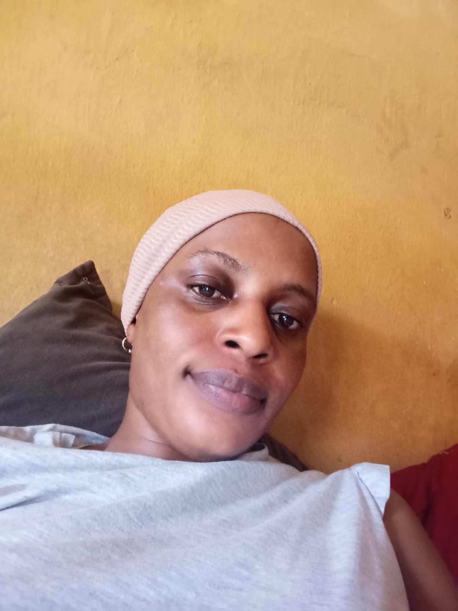 A medium shot shows a dark-skinned woman in a light gray t-shirt and a light tan head covering. She is seated against a brown pillow and a red cushion, positioned in front of a yellow wall. The background is out of focus. No specific details indicate the location as Narayi, Nigeria.