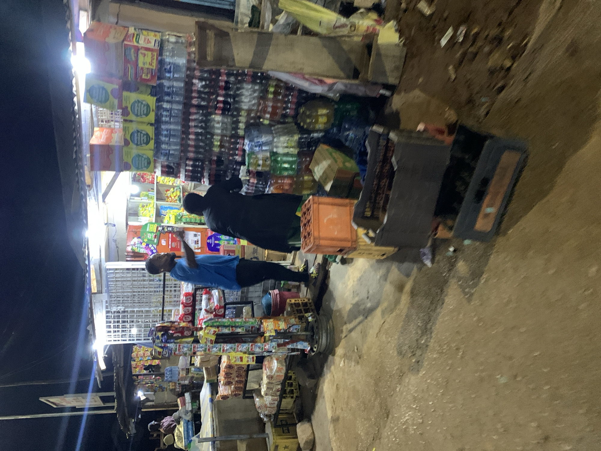 The image shows an outdoor retail space in New Stadium, Nigeria. Two individuals stand near shelves heavily stocked with goods. One person wears a blue shirt and black pants, seemingly reaching up toward the shelves. The other person is dressed in dark clothing. A variety of products are visible, including packaged foods, beverages, and other miscellaneous items. Crates and other storage containers sit on the ground. The surroundings show a dusty, unpaved area. The image is taken at night, illuminated by the bright lights of the shop.