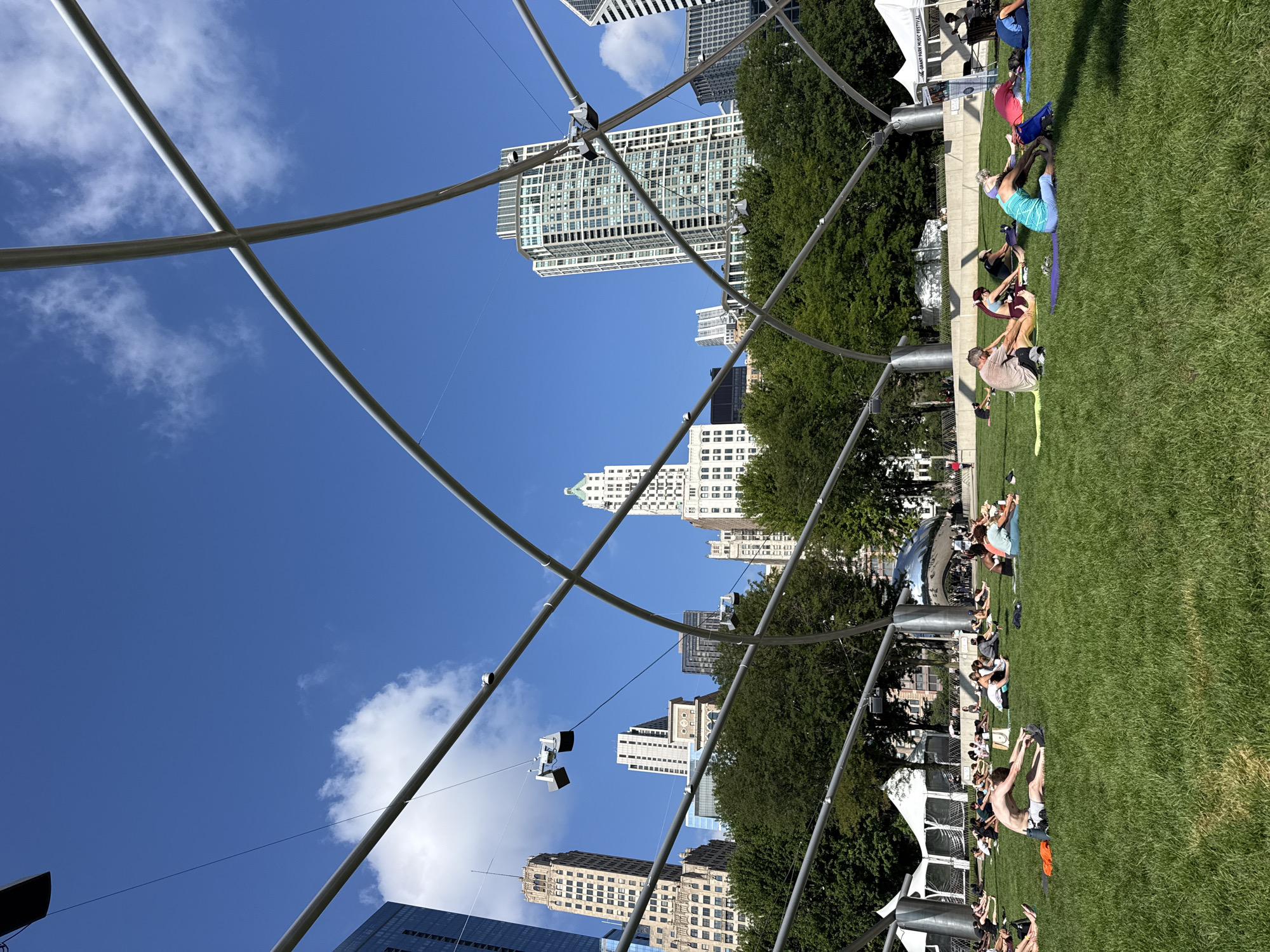 The image captures a vibrant outdoor scene at Millennium Park in Chicago, USA. Under a clear blue sky with scattered white clouds, a group of people are engaged in a yoga or exercise session on a lush green lawn. They are spread out on individual yoga mats, some in seated positions with legs extended, while others are in various stretching poses. The iconic architecture of Chicago serves as a backdrop, with several tall buildings, including skyscrapers with distinct facades, visible above a line of mature trees. A silver, bean-shaped sculpture is partially visible in the mid-ground. Overhead, a large, curved metal structure, likely part of the park's infrastructure, frames the sky. Tents are also present in the background, suggesting a public event or gathering. The atmosphere appears relaxed and communal, with individuals focused on their practice.