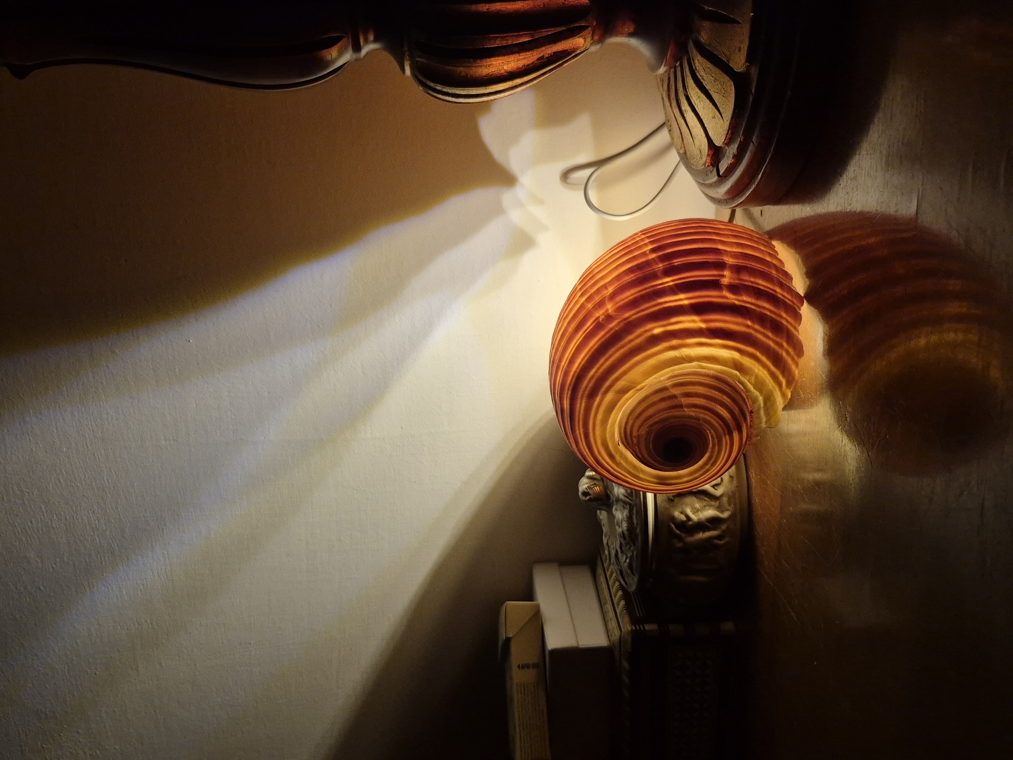 The image shows a close-up of a seashell, lit from within by an unseen light source, casting dramatic shadows and highlights on a textured wall and a wooden surface. The seashell is a spiral shape with translucent orange and yellow bands. To the left of the shell, stacked books and a decorative lidded box are visible, partially obscured by shadow. To the right, the base of a dark, ornate lamp with carved details is present. The lighting suggests a nighttime or dimly lit indoor setting. No people are present, and no specific location in Taranto, Italy, can be identified from the image.