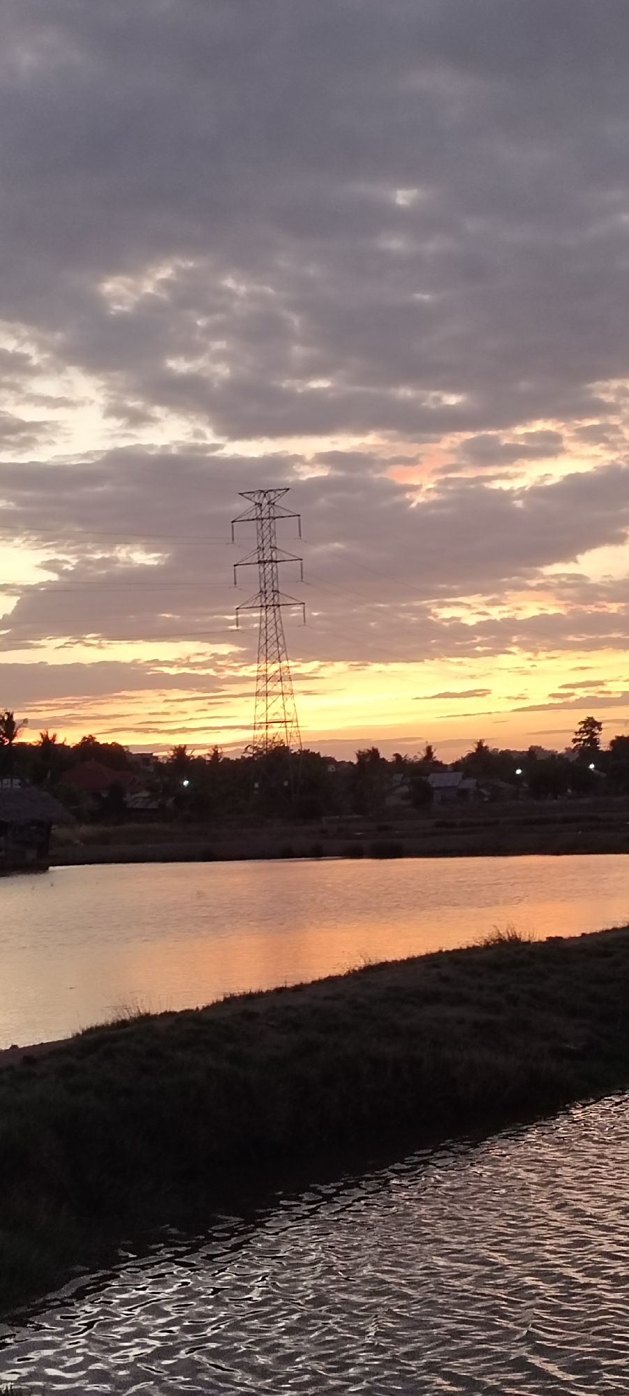 The image depicts a serene sunset scene in Lhokseumawe, Indonesia. The sky is filled with a dramatic display of clouds, transitioning from deep gray at the top to soft oranges and yellows near the horizon. A prominent electricity pylon stands silhouetted against the vibrant sky, a symbol of modern infrastructure juxtaposed with the natural beauty of the sunset. Below, a calm body of water reflects the warm hues of the sky, its surface rippled by a gentle breeze. The shoreline is lined with dark, grassy vegetation, and in the distance, a cluster of houses and trees forms a village. The overall atmosphere is peaceful and tranquil, capturing the ephemeral beauty of twilight in this Indonesian city.
