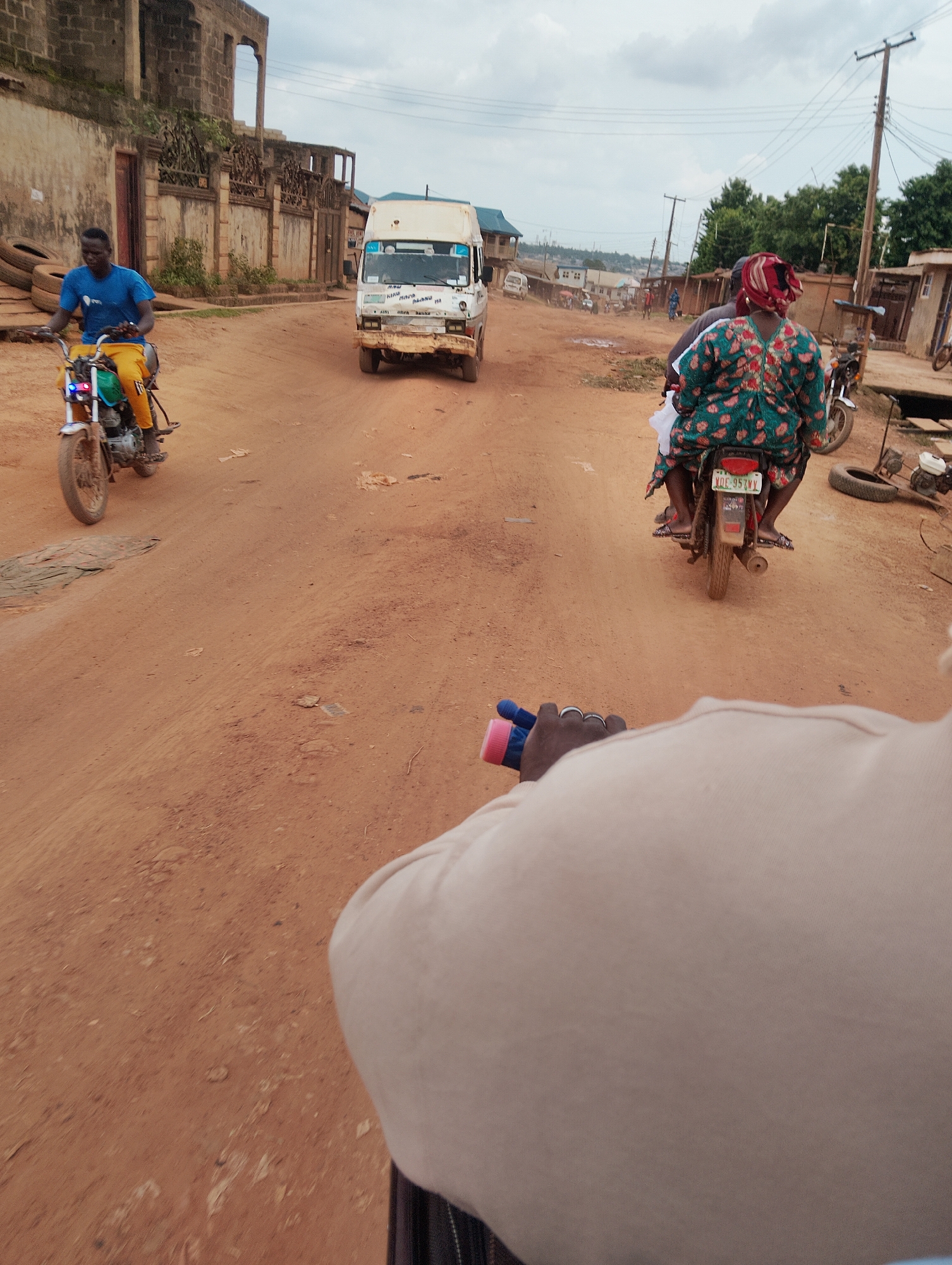 A point-of-view shot taken from the back of a motorcycle on a dusty, unpaved road in Agege, Nigeria. The road is a reddish-brown dirt and there are loose stones scattered on it. In the foreground, a person's hands, wearing rings, are visible holding onto the handlebars of the motorcycle, one hand with a blue and pink object attached.
Further up the road, a man in a blue t-shirt and yellow trousers rides a motorcycle, its front light flashing red and blue. Beside him, a white minibus with writing on its side is driving in the same direction. To the right of the minibus, another motorcycle is visible with two people on it; the passenger in the back wears a patterned green and red dress and a red headscarf. Power lines are visible overhead, supported by wooden poles. In the background, there are buildings with unfinished walls and some trees. The sky is overcast with clouds.