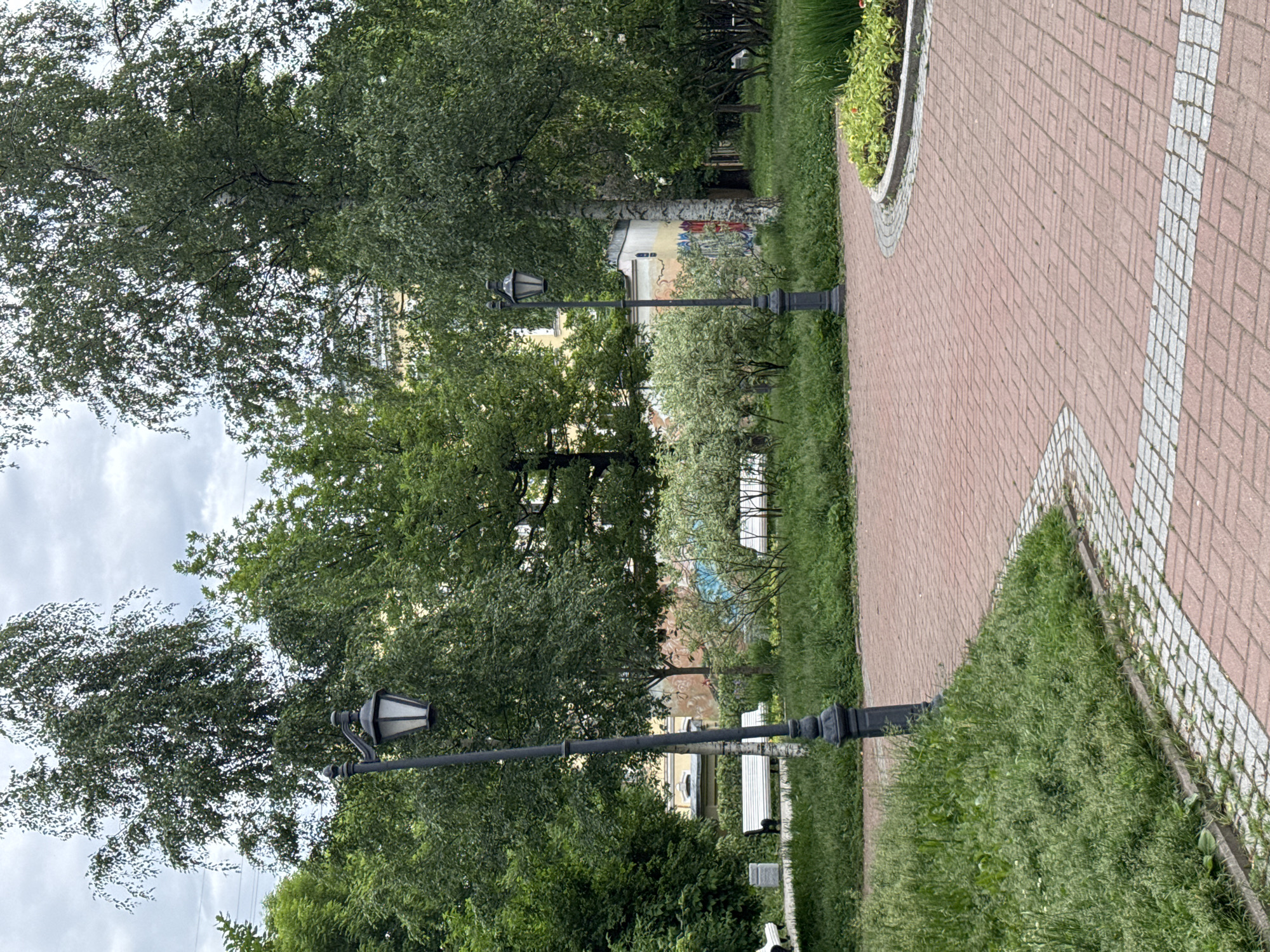 The image depicts a park scene in Saint Petersburg, Russia, on a cloudy day. A red brick pathway with a granite inlay curves through the frame, bordered by lush green grass and trees. Two vintage-style black lampposts stand on either side of the path. In the background, white benches are visible, along with a building adorned with colorful murals. A flower bed with green plants and small orange flowers is situated to the right of the path.
