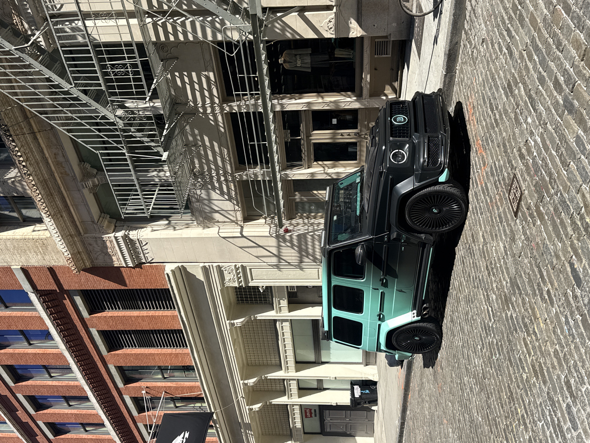 A luxury vehicle is parked on a cobblestone street in a district of New York City, United States. The vehicle is a dark-colored SUV with distinctive styling, including blacked-out wheels and an unusual light green accent on the lower portion of the vehicle, which is an otherwise black vehicle. The vehicle is parked along the curb of a street lined with multi-story buildings with ornate facades. A fire escape is affixed to the exterior of one building. The buildings appear to house both retail and residential spaces. The image is oriented at an unusual angle, with the top of the buildings tilted to the left, suggesting the photograph was taken from an askew perspective. No people are visible.