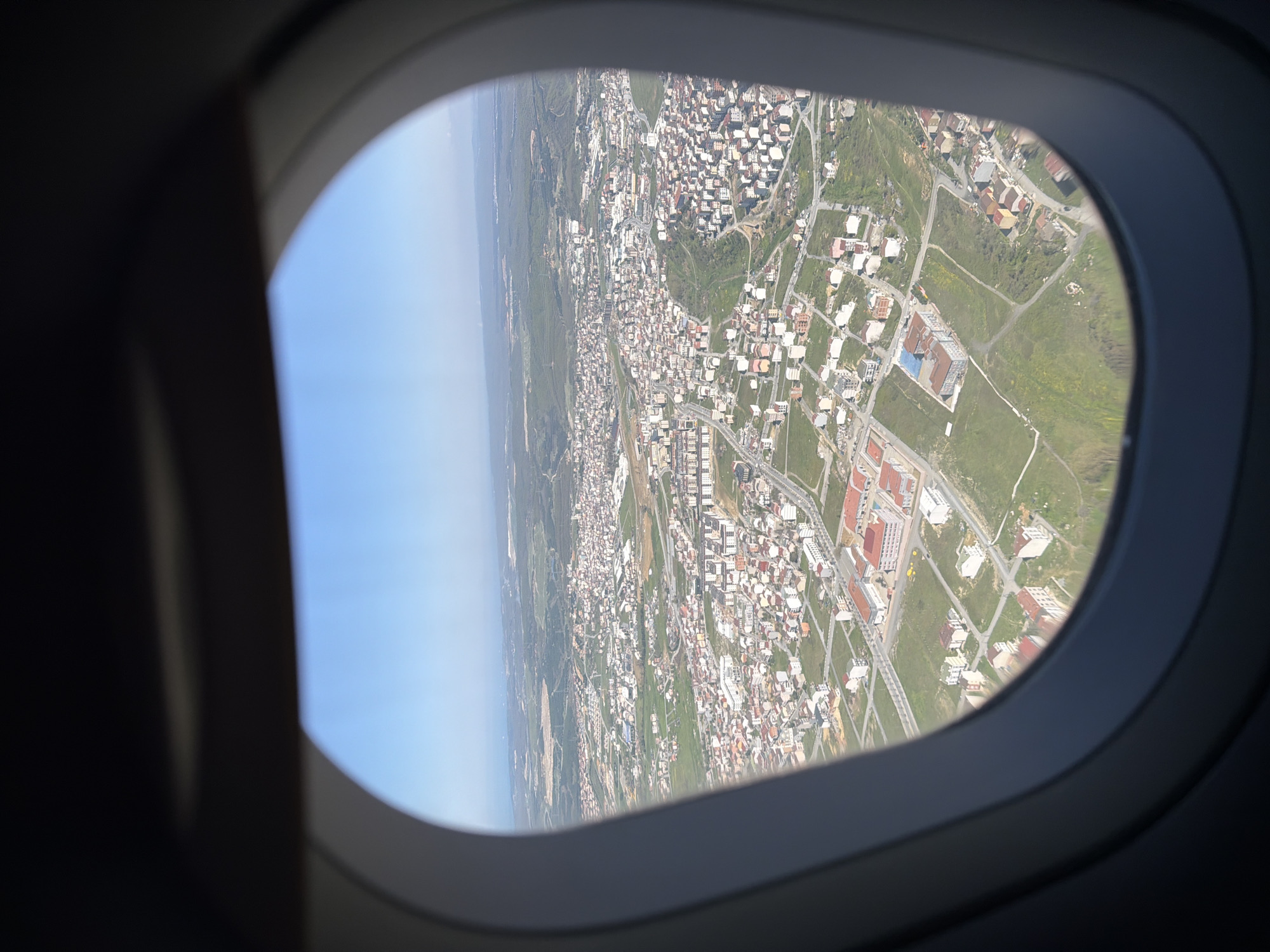 The image provides an aerial view of a populated area, seen through the dark gray, oval-shaped window frame of an airplane. The scene outside the window is bathed in bright daylight under a clear blue sky, suggesting good weather conditions.
The landscape below features a dense arrangement of buildings, primarily light-colored residences with a variety of roof types, including some with red or orange tiles. These structures are interspersed with green patches of land, indicating a mix of developed and natural areas, possibly suburban or a town bordering undeveloped land. Roads are visible, winding through the settlements. In the far distance, a distinct horizon line separates the land from the clear sky, potentially indicating a coastline or a large body of water.
The photograph appears to have been taken with the camera rotated 90 degrees clockwise, as the horizon line runs vertically along the left edge of the visible landscape through the window. No people or visible text are present in the image. The scene captures the experience of looking down upon a community from an aircraft during flight.