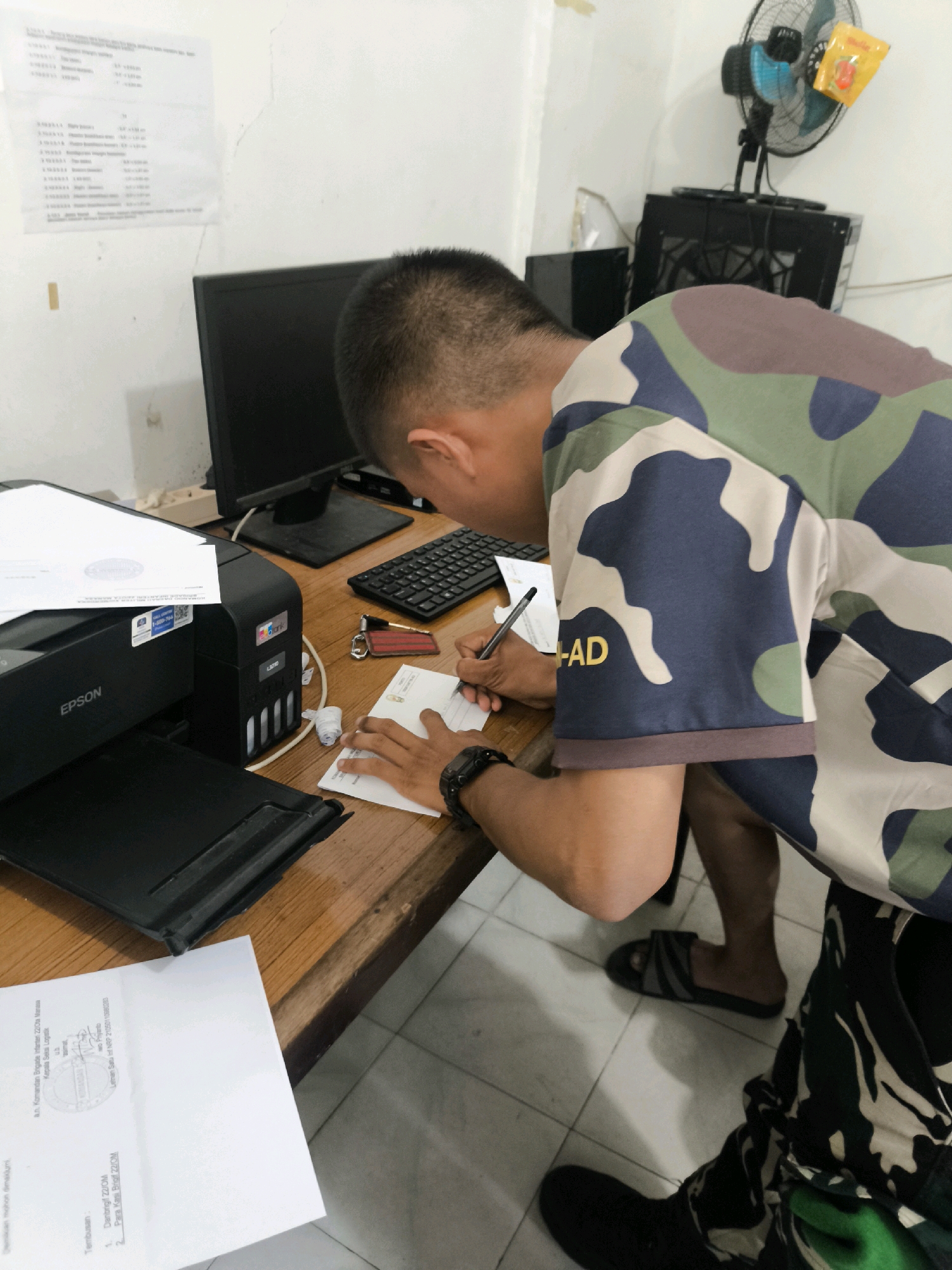 A young man wearing a camouflage shirt is seated at a wooden desk, writing on a piece of paper. To his left, an Epson printer sits on the desk, with papers on top of it. A computer monitor and keyboard are also visible on the desk. The man is wearing sandals and his pants are also camouflage. In the background, a fan is mounted on the wall, and a rack of computer equipment is behind it. The scene appears to be an office or a workspace.