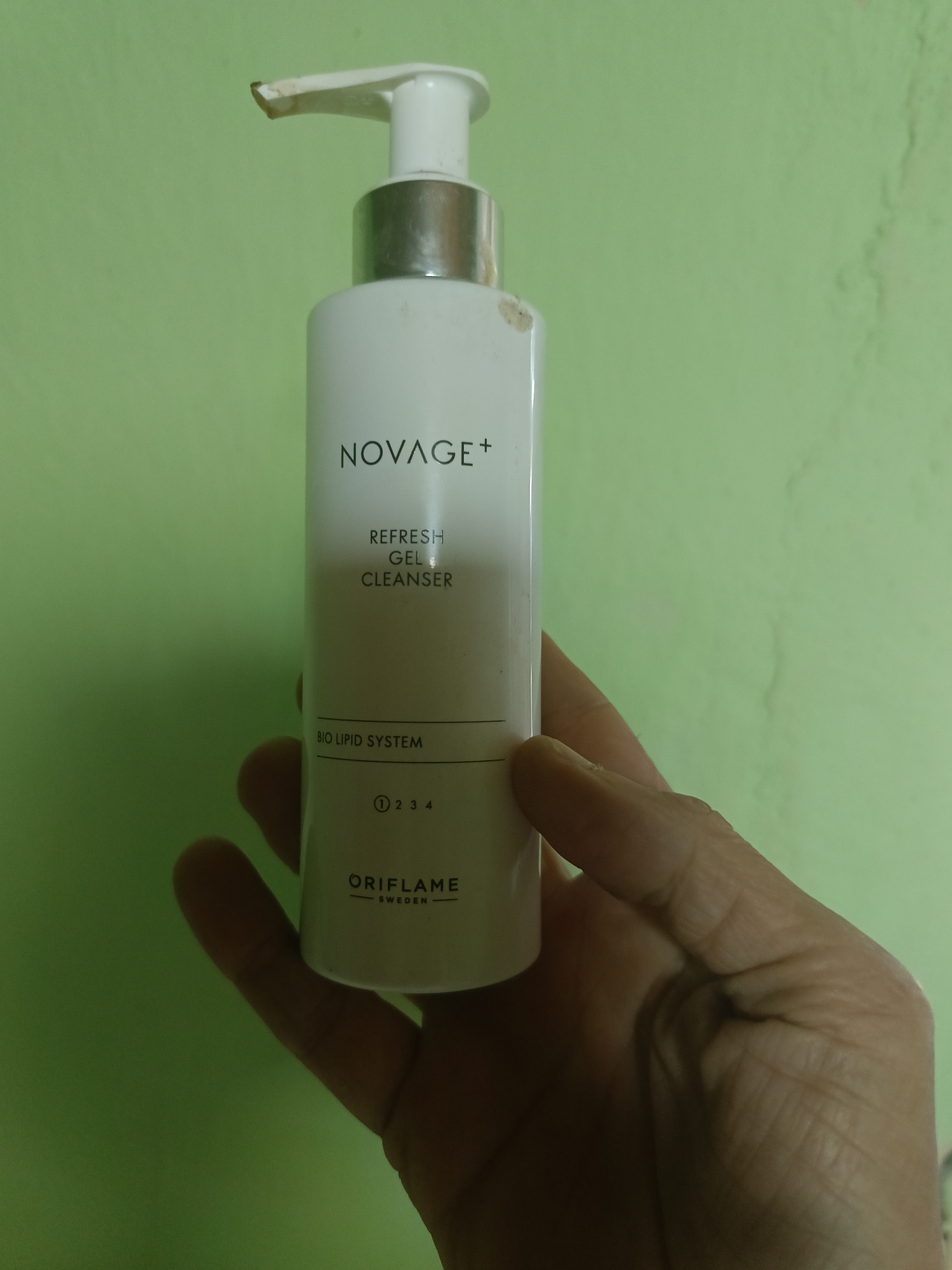 A white pump bottle of "NOVAGE+ REFRESH GEL CLEANSER" is held by a person's hand against a pale green, textured wall. The bottle has silver accents around the pump and neck. The label features the brand name "NOVAGE+" prominently, followed by "REFRESH GEL CLEANSER". Below that, "BIO LIPID SYSTEM" is written, and then a series of numbered circles "1 2 3 4". At the bottom of the visible label, "ORIFLAME SWEDEN" is printed. There are some minor smudges on the pump and bottle. The background is a plain, light green wall with some subtle discoloration or texture variations. The scene is indoors.