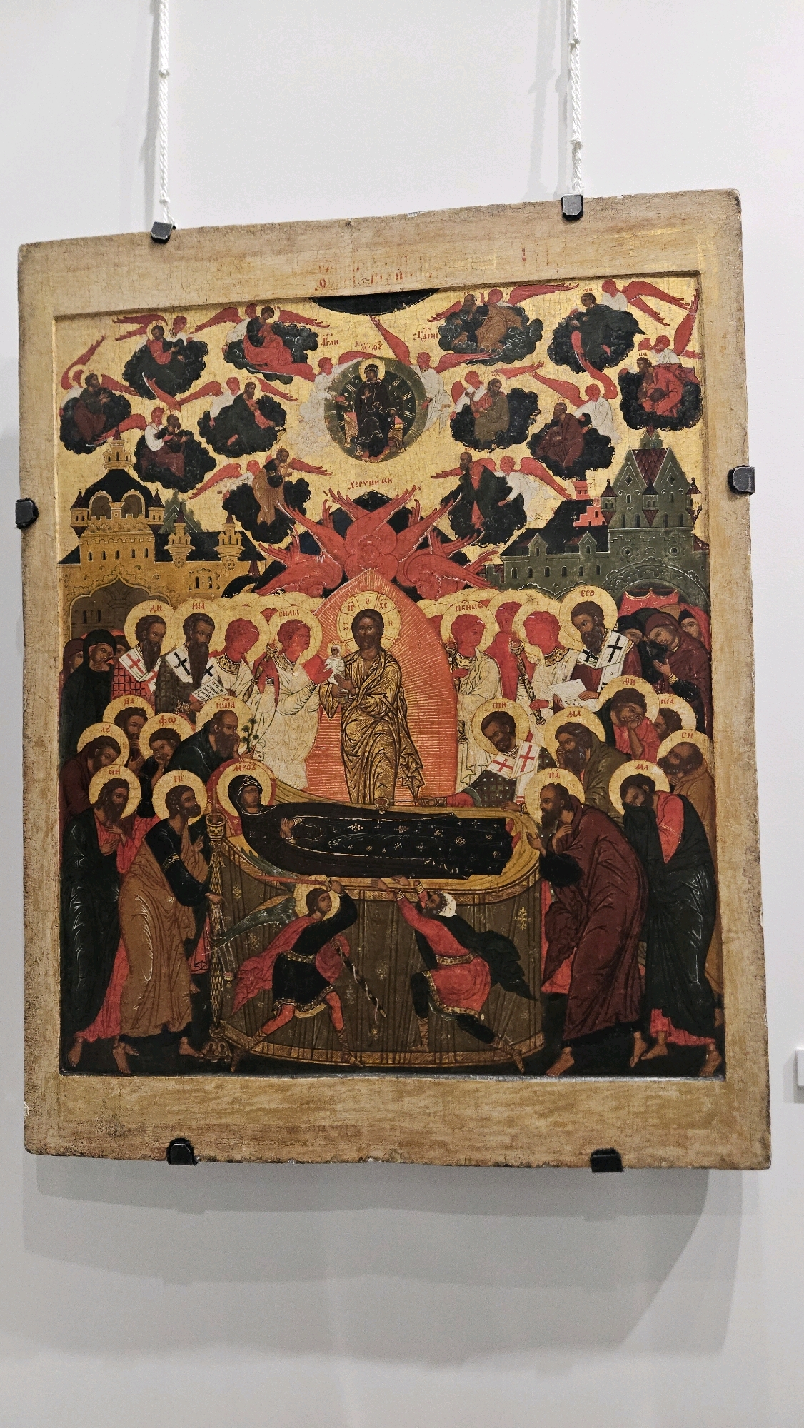 This is an image of an icon depicting the Dormition of the Theotokos. The scene is set against a golden background with architectural elements and numerous angelic figures in the upper register. At the center of the lower portion of the icon, the Virgin Mary lies on a funeral bier, appearing to be asleep. Christ is shown in the upper center, receiving her soul, depicted as a child. Surrounding the bier are the Apostles and other disciples, some of whom are looking at Mary, while others are engaged in prayer or lamentation. There are also figures depicted as a threat to the bier, being struck down. The icon is framed by a wooden border and appears to be hanging on a white wall, possibly in a museum or church setting.
While the image itself does not provide specific location details, this icon is part of the collection of the Ulyanovsk Regional Art Museum in Ulyanovsk, Russia.