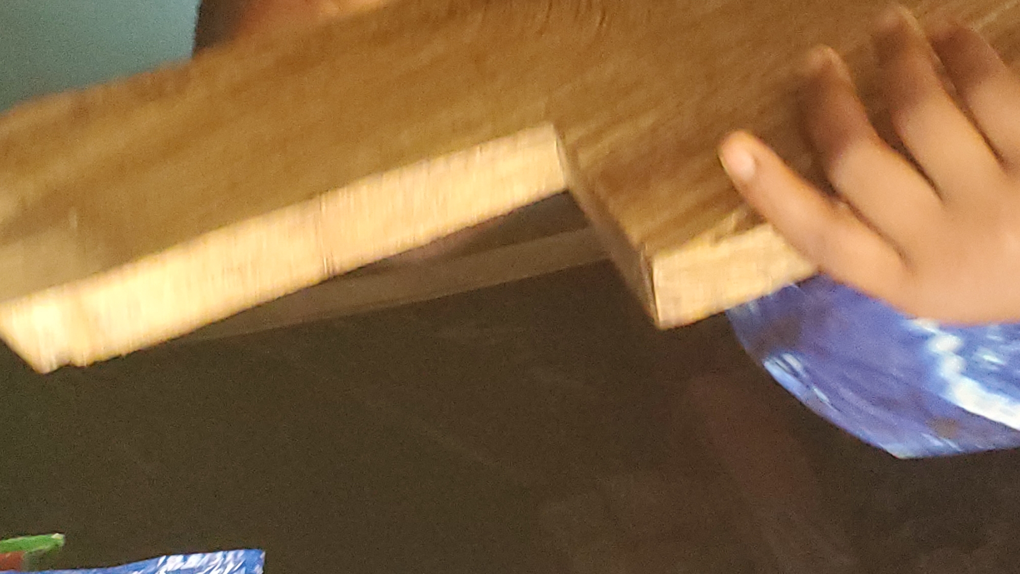 This media file shows a close-up of a person's hand holding a wooden plank. The plank is light brown and appears to be made of pine or a similar softwood. The hand is child-sized, with dark skin. The person is wearing a blue and white tie-dye garment, possibly a shirt or a wrap. The background is blurred and dark, suggesting an indoor setting with low lighting. There are some indistinct shapes and colors in the lower part of the frame, which might be other objects or materials. The image is focused on the interaction between the hand and the wood, highlighting the texture and grain of the material. There is no visible text in the image. The setting is likely Jalingo, Nigeria, but there are no specific visual cues to confirm this. The overall impression is one of a simple, everyday moment, possibly related to construction, crafting, or play.