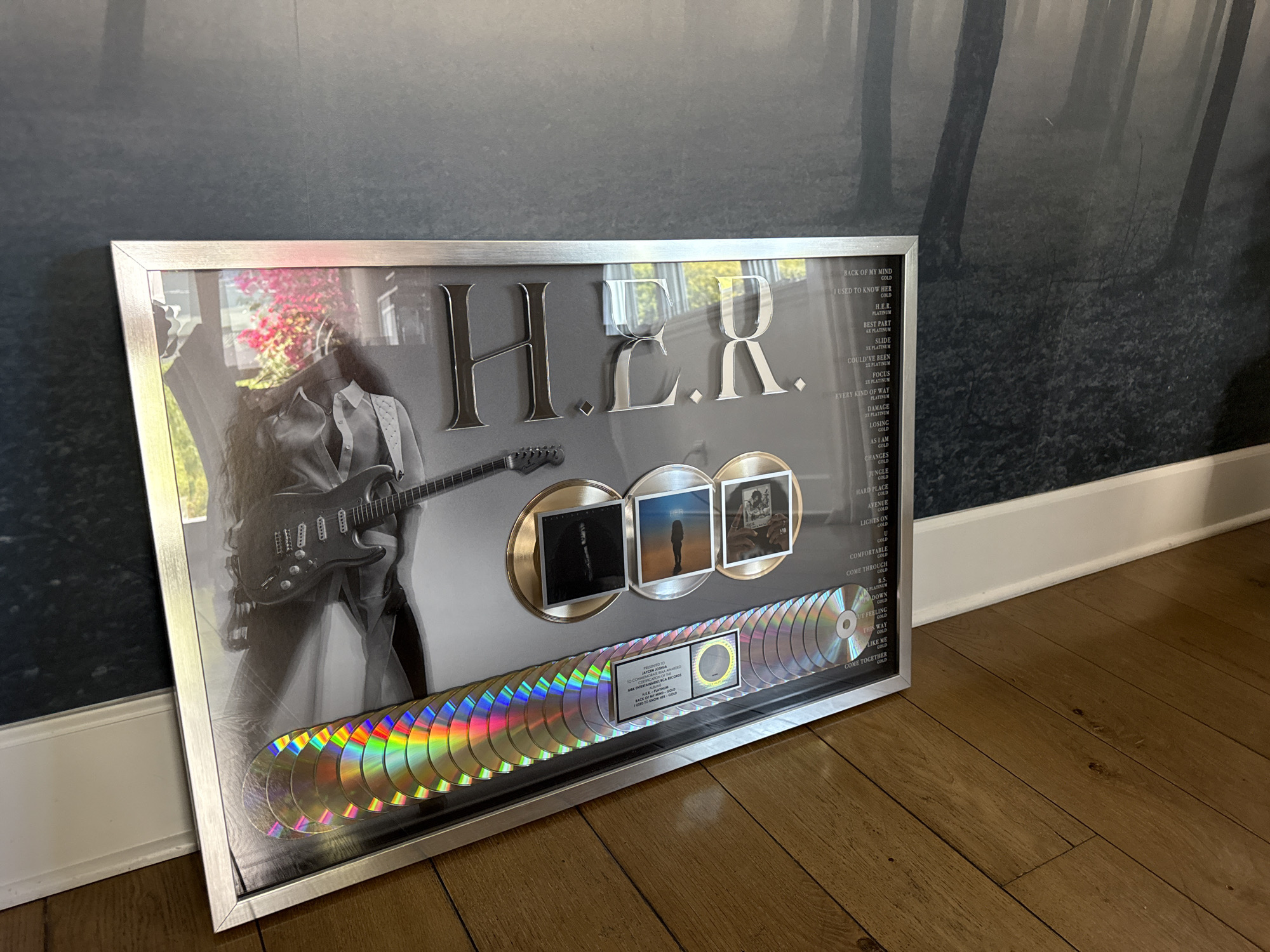 Here's a description of the image:
The image shows an award commemorating H.E.R.'s musical achievements, presented to Jaycen Joshua, possibly in a home setting given the wood flooring and textured wall behind it. The award features a black-and-white photo of H.E.R. playing guitar, the name "H.E.R." in large silver letters, three album covers mounted on gold discs, and a row of what appears to be compact discs at the bottom. A plaque lists H.E.R.'s songs and their platinum/gold status, signifying their sales achievements.