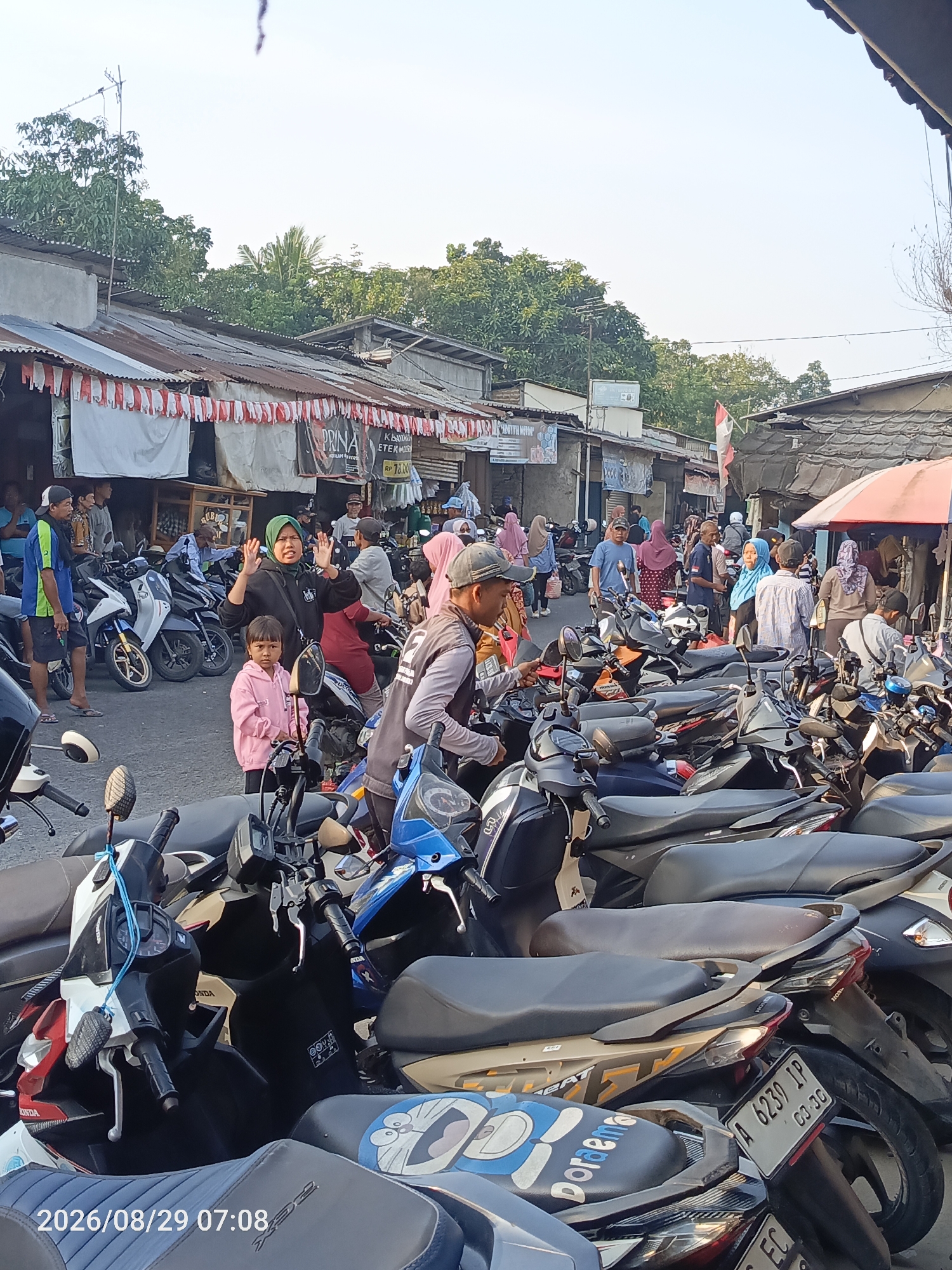 The image depicts a bustling street scene in Sangiang, Indonesia, likely a marketplace or a busy thoroughfare on a clear day. The foreground is dominated by a dense arrangement of parked motorcycles, indicating a significant amount of vehicular traffic. A number of people are visible, some interacting with each other, while others are standing or walking amongst the parked vehicles.
In the midground, a woman wearing a green hijab and a dark jacket is gesturing with her hands, seemingly in conversation or addressing someone. A young child in a pink hoodie stands beside her. Behind them, a crowd of people are milling about, some carrying shopping bags, suggesting a commercial area.
The background reveals a row of shops and stalls with corrugated iron roofs, many adorned with red and white Indonesian flags, hinting at a national celebration or patriotic decoration. Trees with dense foliage line the street behind the buildings, providing a natural backdrop to the urban environment. The sky is bright and clear, suggesting daytime with good weather.
The overall impression is one of a lively, populated area in Indonesia, characterized by motorcycle transportation and active commerce. The presence of flags suggests a possible festive atmosphere or a sense of national pride.