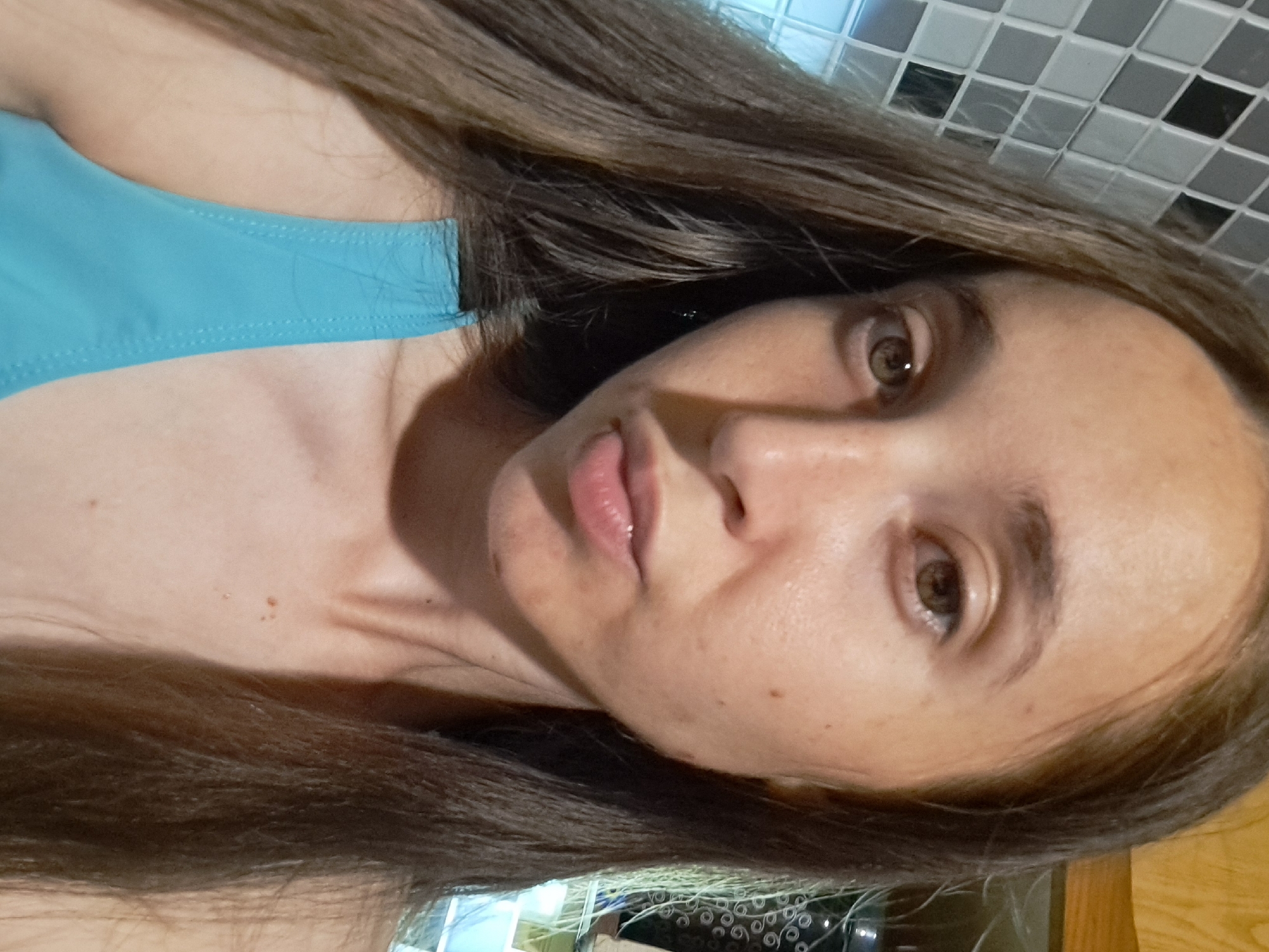 This is a close-up, eye-level, selfie-style photograph of a young woman with long, brown hair. She is wearing a turquoise swimsuit top. Her face is the primary subject, with her head slightly tilted. The background shows a tiled wall with a diamond pattern and what appears to be a wooden surface to the right, possibly a cabinet or shelf. There are also some indistinct shapes and lighting behind her. The woman's skin shows some freckles and minor blemishes. The photograph is taken indoors. The location of Vypasne, Ukraine is not visually evident in the image.