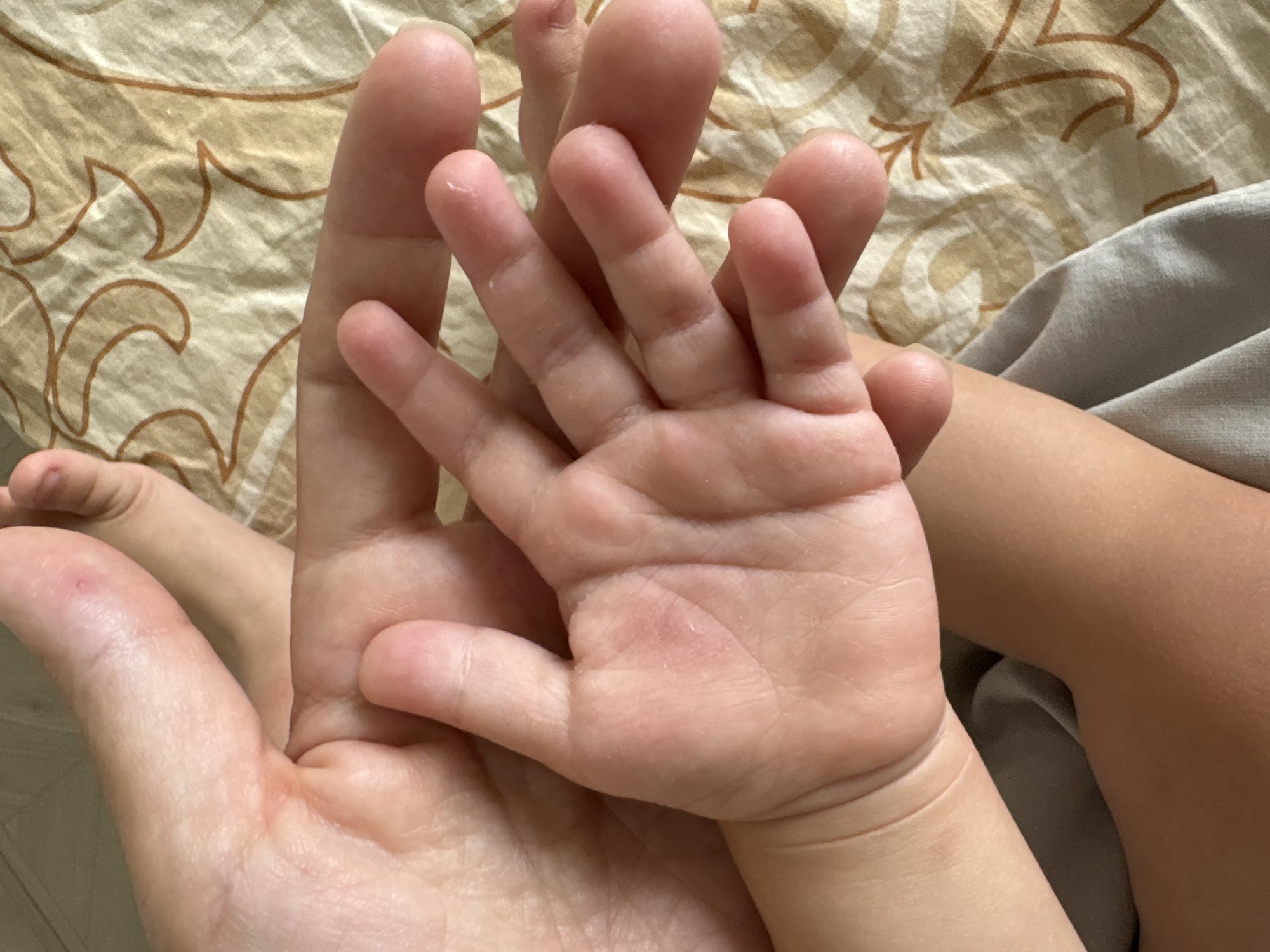 The image shows an adult hand holding a child's hand. The child's hand is smaller and placed on top of the adult hand. Both hands are pale and pinkish in color. The background is a bed with patterned bedding. The scene appears to be indoors and possibly residential. The location is Ембаево, Russia.