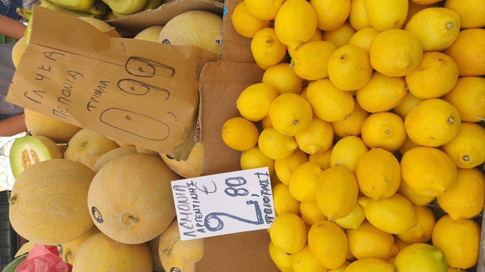 A street fruit stall in Neoi Epivates, Greece, displays a variety of produce. In the foreground, a large pile of bright yellow lemons is visible, with a white sign indicating a price of "2,80 €" and "ΠΡΩΤΗ". Behind the lemons, several cantaloupes are stacked, one of which is cut in half, revealing its green flesh. A brown paper bag with hand-drawn markings and writing in Greek, including "ΓΛΥΚΑ ΠΕΠΟΝΙΑ" and "ΤΡΙΑΛΑ", is also present, with "0,99" written prominently, suggesting a price. A person's hands are partially visible in the upper right corner, reaching towards a box of bananas. The background shows additional fruit and what appears to be a market environment.