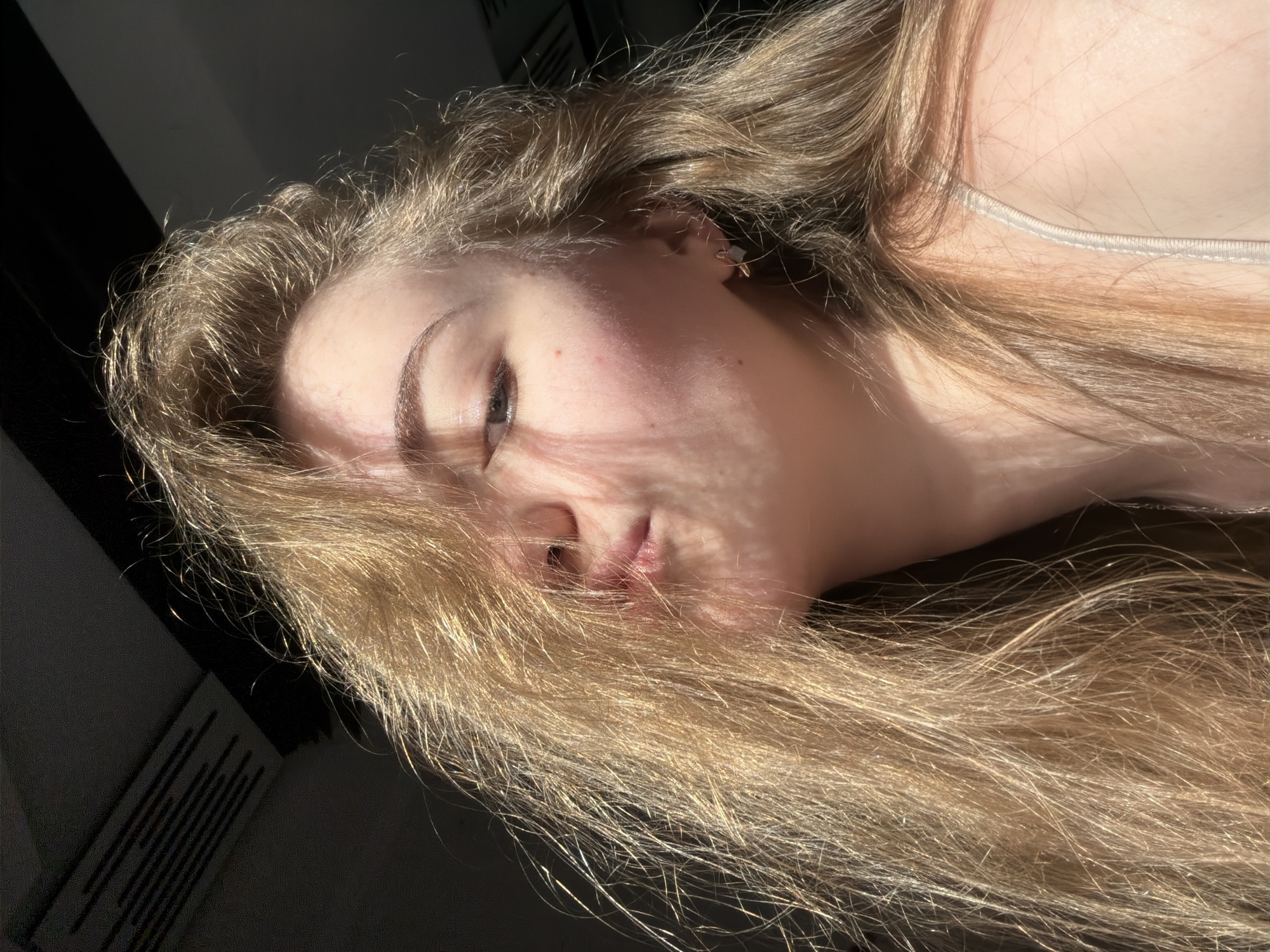 An extreme close-up, selfie-style portrait of a young woman with long, blonde hair partially obscuring her face. Her head is tilted, and she is puckering her lips. Sunlight streams across her face, casting shadows and highlighting individual strands of hair. She is wearing a thin, light-colored strap, likely from a camisole or bra, across her shoulder. The background is dark and out of focus, suggesting an indoor setting. There are no discernible landmarks or environmental cues to suggest a specific location within the city of Dnipro, Ukraine.