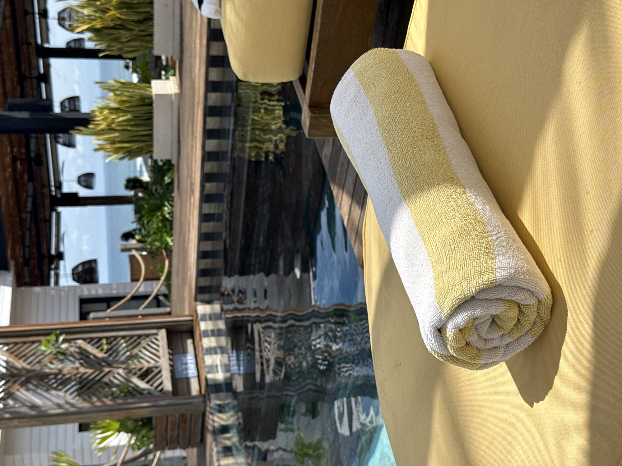 A close-up shot showcases a rolled yellow and white striped towel on a yellow lounge chair. The composition captures the texture of the towel and the lounge chair. In the blurred background, there is a pool reflecting the surrounding buildings, foliage, and the sky. The location is identified as North Kuta, Indonesia.