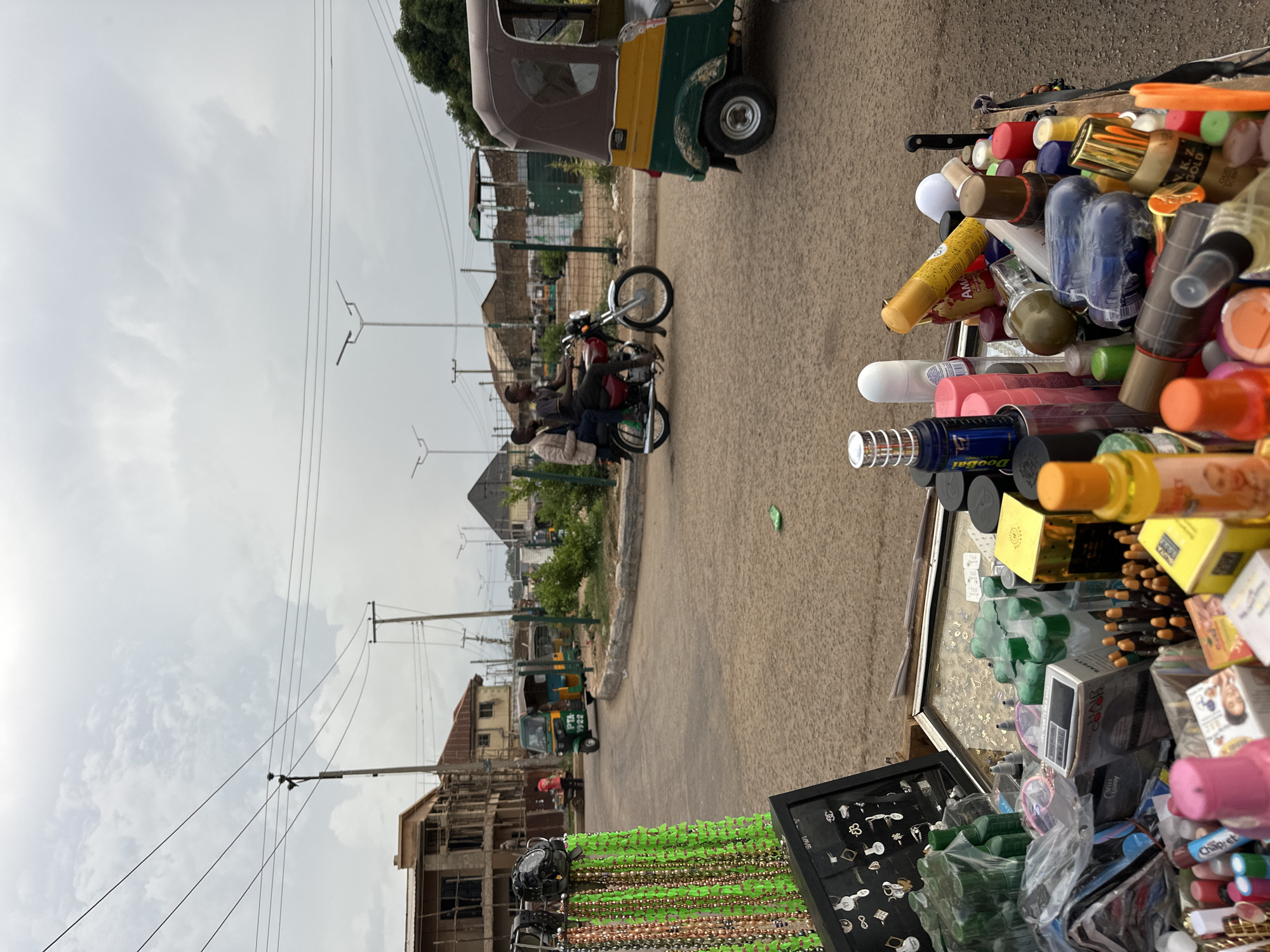 In Bukuru, Nigeria, a street vendor's stall displays an assortment of goods, including toiletries and accessories. Among the items are hair color products, perfumes, body sprays, and jewelry. In the background, a busy intersection shows traffic with a motorcycle carrying two individuals, and several auto-rickshaws, known as Keke Napeps. Construction is underway on a multi-story building to the left, and utility poles with overhead wires are visible. The sky is overcast.
