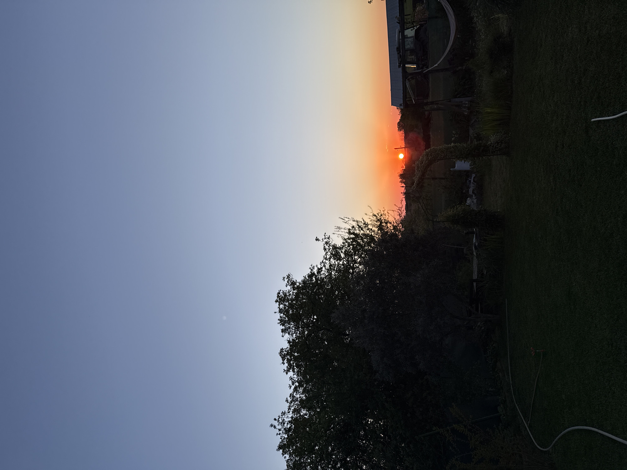 The image shows an outdoor scene in Generalovo, Bulgaria, at dusk or dawn. The sky transitions from blue to orange around the horizon, where the sun is visible. Below the horizon, there are trees and vegetation, including a small archway covered in plants. To the right, there is a patch of green grass. In the background, there appears to be a building or structure. A garden hose is visible in the right of the image on the ground.