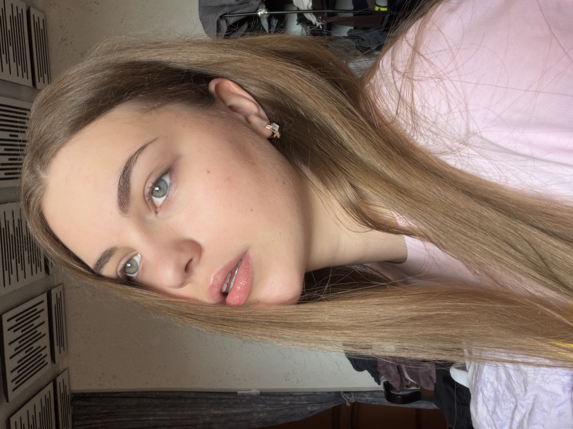 A young woman with long, blonde hair and light blue eyes is shown from the chest up, looking upwards and to the left. She is wearing a light pink t-shirt and a small, square earring in her right ear. The background is out of focus and appears to be an indoor setting with what looks like acoustic panels on the ceiling and a curtain on the left. There is a shelf with clothing items visible on the right side of the image. The lighting suggests it is daytime. The location is specified as Dnipro, Ukraine.