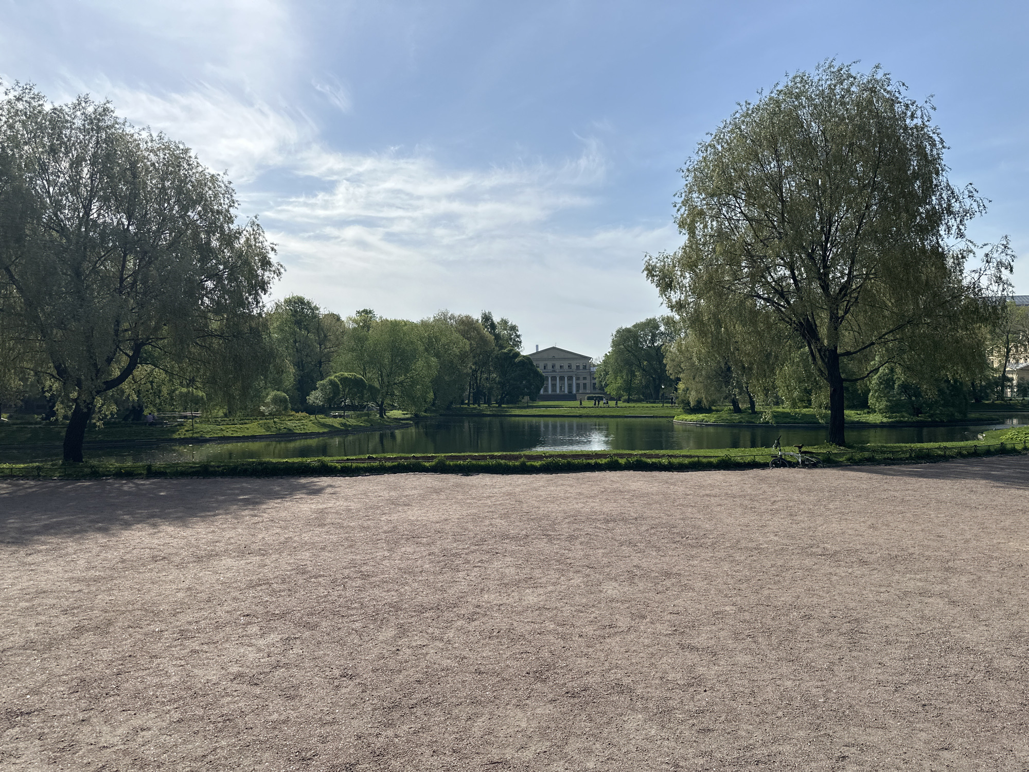 The image depicts a park setting in Saint Petersburg, Russia. A gravel area in the foreground leads to a body of water, likely a pond or small lake, bordered by green grass and shrubs. Trees frame both sides of the view. In the distance, a building with columns is visible. The sky is blue with wispy clouds. There are no people visible in the image. A bicycle is leaning against a tree on the right-hand side of the water.