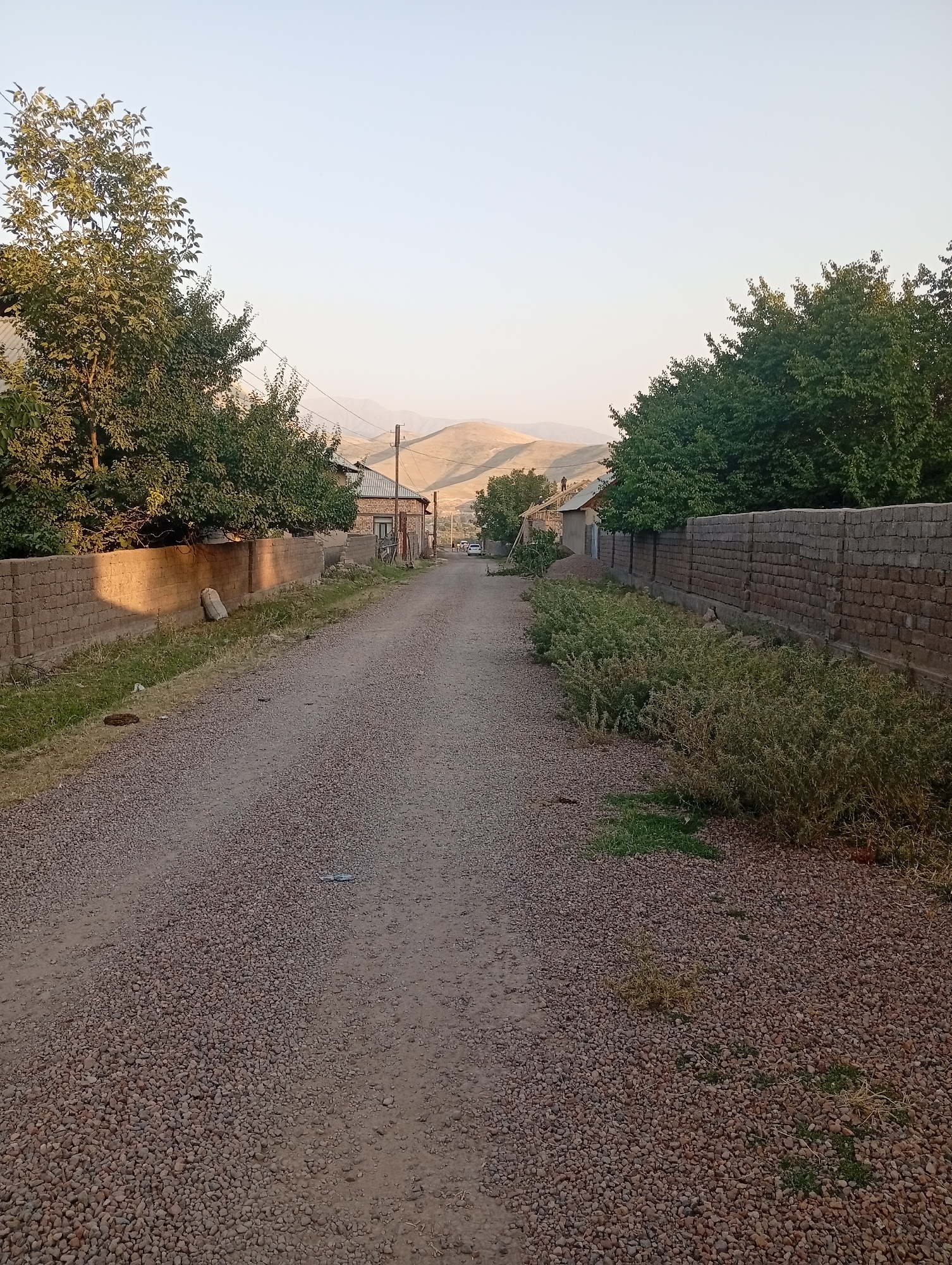 The image depicts a gravel road in a rural area, likely a village within the city of Angren, Uzbekistan. The road is flanked by low brick walls and dense green vegetation on either side. On the left, a brick wall casts a long shadow, indicating late afternoon or early morning light. A sandbag rests against this wall. Trees with lush green foliage are visible on the left and right sides of the road, partially obscuring buildings. In the distance, rolling hills dominate the horizon under a pale, clear sky. Several houses with brick facades and tiled roofs are scattered along the road. Some appear to be under construction, with visible roof framing. A white car is parked on the right side of the road further up. The overall scene suggests a quiet, residential environment in a hilly, arid landscape.