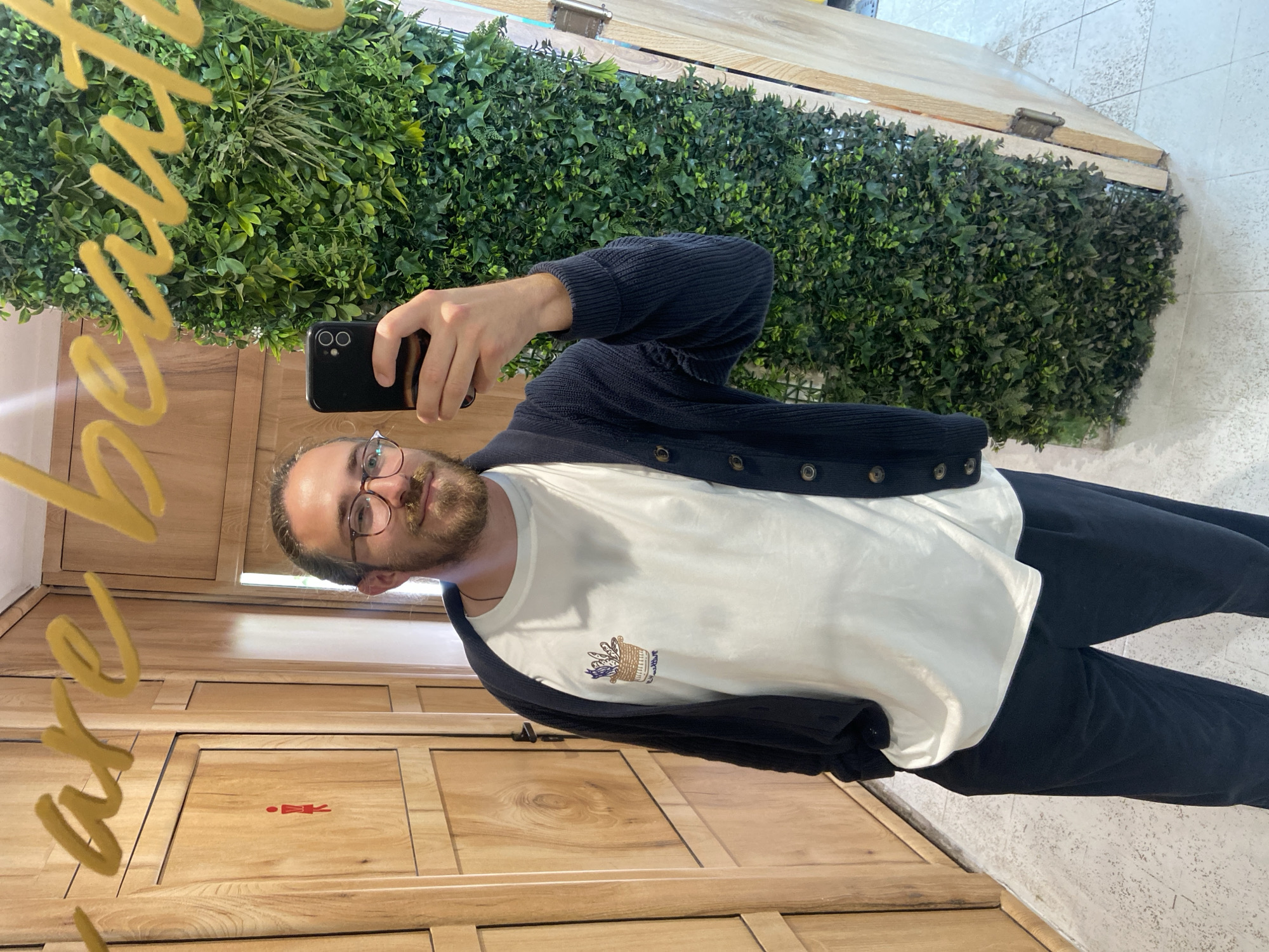 A man is taking a selfie in what appears to be a restroom or changing area. He is holding a black smartphone in his right hand. The man is wearing glasses, a white T-shirt with a small emblem on the chest, a dark blue cardigan, and dark trousers.
The backdrop includes a wall covered with artificial greenery. A mirror is partially visible, reflecting the man and a gold-colored text design. The restroom features wooden-paneled walls and doors, with a pictogram indicating a female restroom.
While Petrovac centar, Montenegro, is specified as the location, there is no conclusive visual evidence in the image to independently confirm this.