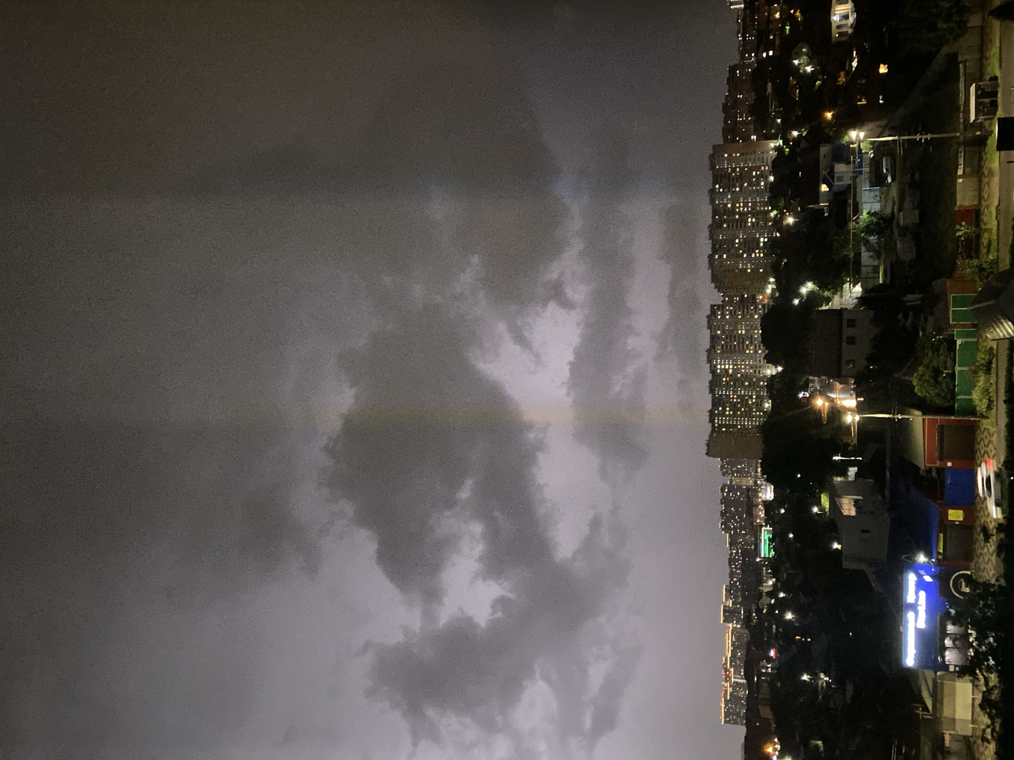 The image captures a nighttime city skyline under a cloudy sky, likely during or after a storm. The sky is filled with dark, heavy clouds, with a faint vertical streak of light that could be a rainbow or a light anomaly. Below the sky, numerous city buildings are visible, many of them illuminated with countless lit windows, suggesting residential or office spaces. The city appears to be built on varied terrain, with some buildings situated on higher ground. The foreground shows a cluster of smaller buildings, including houses and what appear to be commercial establishments, some with illuminated signage. Streetlights dot the landscape, casting pools of light on the streets and surrounding areas. The overall scene conveys a sense of urban life at night, with a dramatic sky overhead.