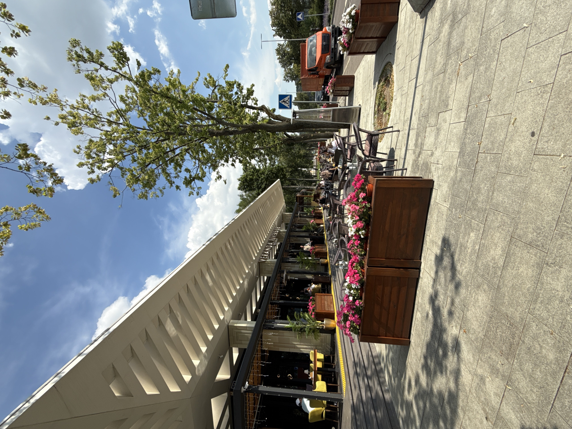 The image depicts an outdoor seating area of a restaurant or cafe, likely in an urban or suburban environment. The scene is oriented vertically, giving a unique perspective. In the foreground, a modern building with a white, slatted roof extends into the frame. Beside the building, there are rows of tables and chairs, some occupied by people. The seating area is adorned with wooden planters filled with vibrant pink and white flowers, adding a touch of color.
Further into the scene, a tree with lush green foliage provides shade. Beyond the tree, a street is visible with an orange truck, a pedestrian crossing sign, and other vehicles. The ground is paved with light-colored tiles, and there are shadows cast by the tree, suggesting it is daytime. The sky is blue with scattered white clouds, indicating pleasant weather.
Visible text includes "VERANDA RIVERSIDE" written on the building's exterior, identifying the establishment. The overall atmosphere appears relaxed and leisurely, with people enjoying the outdoor ambiance. The time of day seems to be late morning or afternoon, given the lighting and shadows.