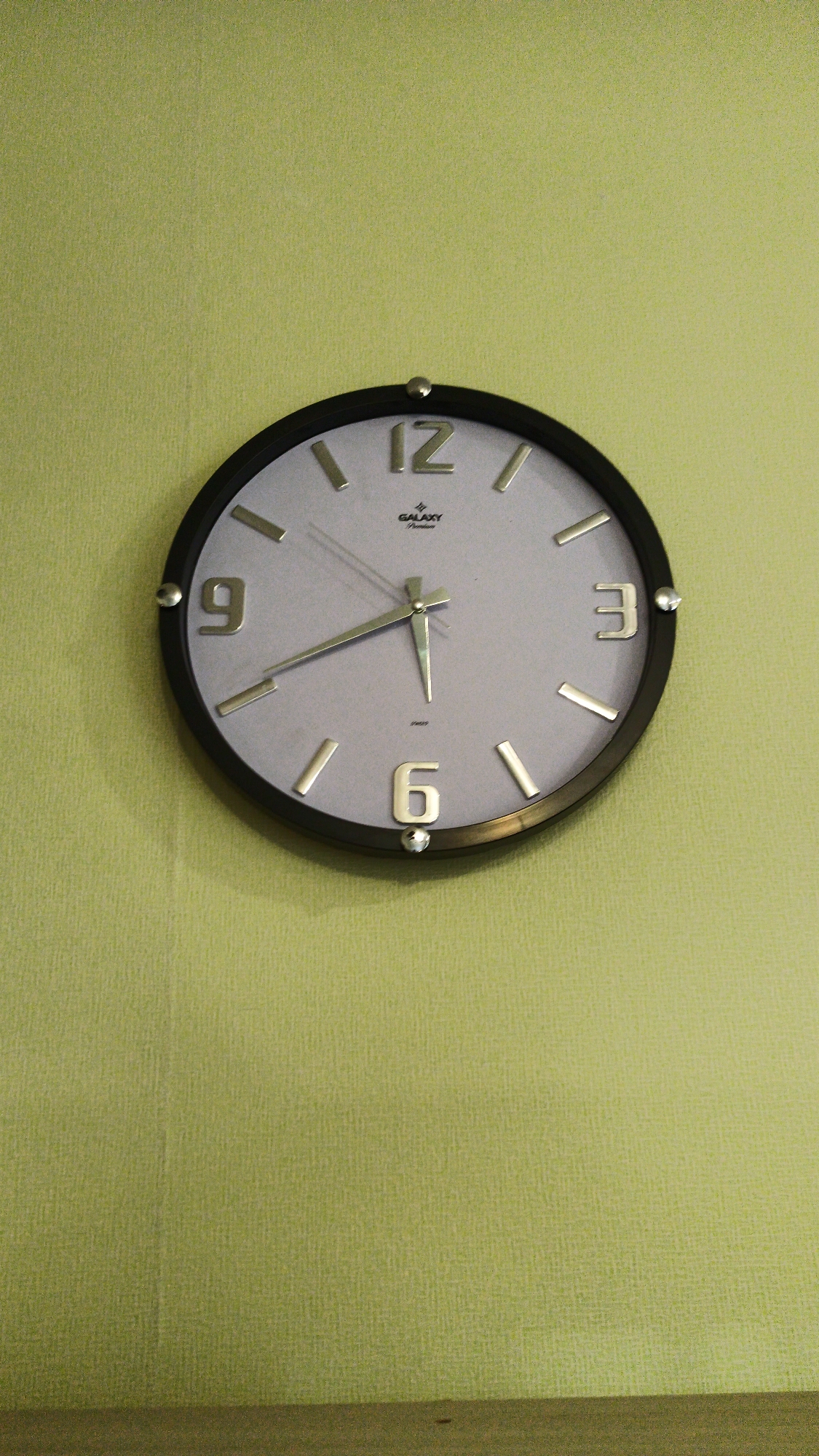 A light gray analog wall clock with a black frame is hanging on a light green textured wall. The clock has silver hour markers and silver numbers at the 12, 3, 6, and 9 positions. The clock hands are also silver. The clock has the brand name "GALAXY Premium" printed below the 12. The wall is a uniform light green with a subtle textured pattern. The bottom of the image shows a portion of a horizontal wooden surface. There are no people visible in the image. The photograph appears to be taken indoors. The location in the city of Abaza, Russia is unknown from the image alone.