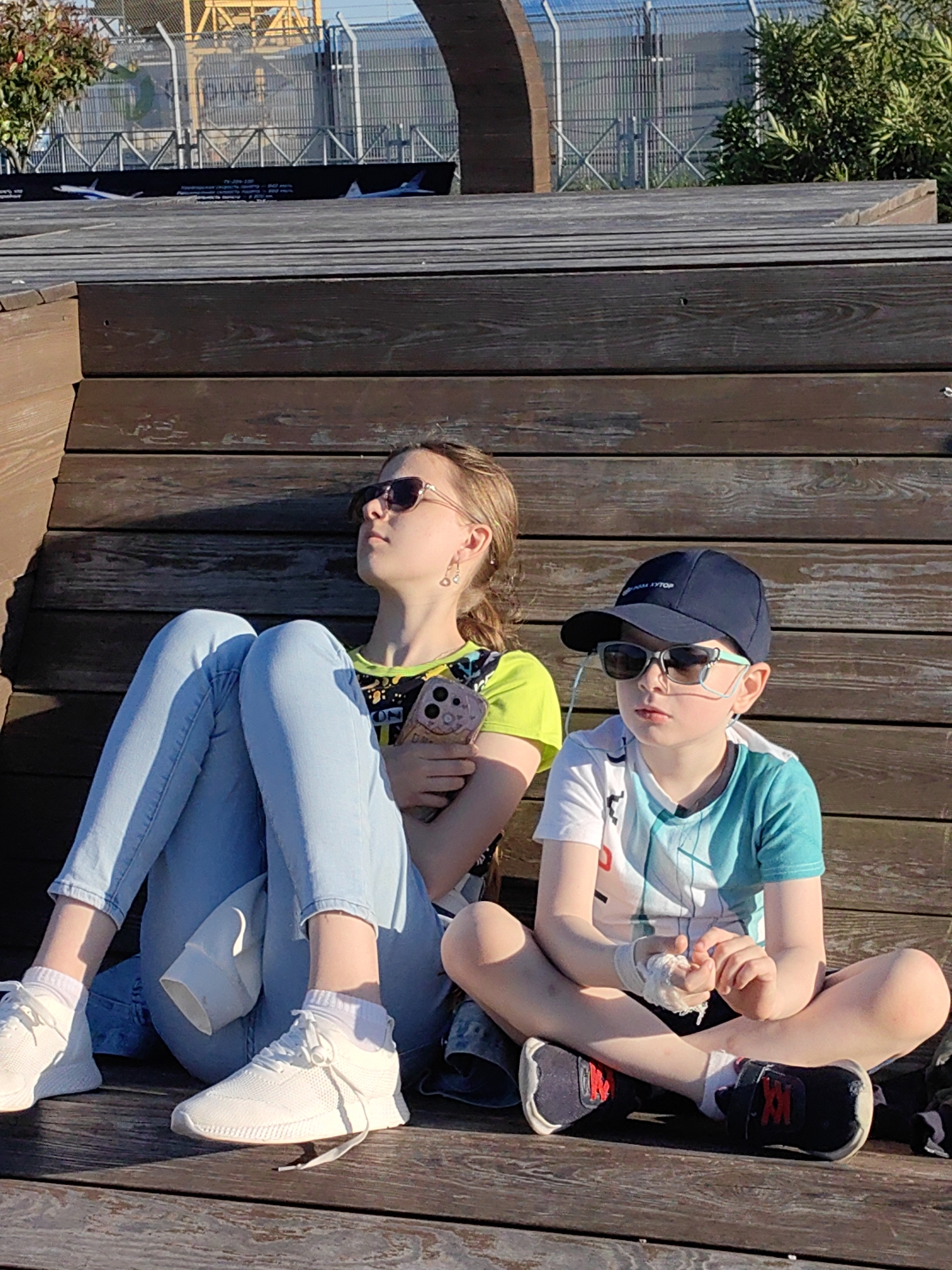 This outdoor image captures two children, an older girl and a younger boy, sitting on a multi-level wooden bench or decking under late afternoon sunlight. They appear relaxed, facing slightly towards the right and looking upwards or into the distance.
The girl, on the left, wears light blue jeans, a bright green and black patterned t-shirt, and white sneakers with white socks. She has her light brown hair pulled back, sunglasses on her face, and small heart-shaped earrings. Her legs are crossed, and she holds a smartphone with a textured pink case in her lap. Her expression is contemplative, with her head tilted slightly up, seemingly enjoying the sun.
The boy, on the right, wears a dark blue baseball cap, sunglasses, a white and teal short-sleeved t-shirt, and dark sneakers with red accents and white socks. His left hand, visible on his lap, appears to have a white bandage or wrap. He sits with his legs crossed, looking straight ahead or slightly to the right, with a thoughtful or observant expression. A thin light blue wire, possibly from an earbud or a medical device, is visible around his ears.
The setting is an open public space with a dark wooden structure forming the seating. In the background, beyond the wooden structure, there is a tall chain-link fence with a large yellow industrial crane or structure behind it. Green foliage is visible on both sides of the fence. A notable detail in the mid-background is a long black banner or billboard placed along the fence line. This banner features silhouettes of airplanes and legible text. The text includes "ПРО АН-124" (About An-124), "скорость полёта" (flight speed), and "дальность полёта" (flight range), strongly indicating an aviation theme, possibly near an airport or an exhibition focused on aircraft.
The sun is low in the sky, casting long shadows across the wooden surfaces, suggesting either late afternoon or early evening. The weather appears clear and sunny. Both children seem to be resting or observing their surroundings.
