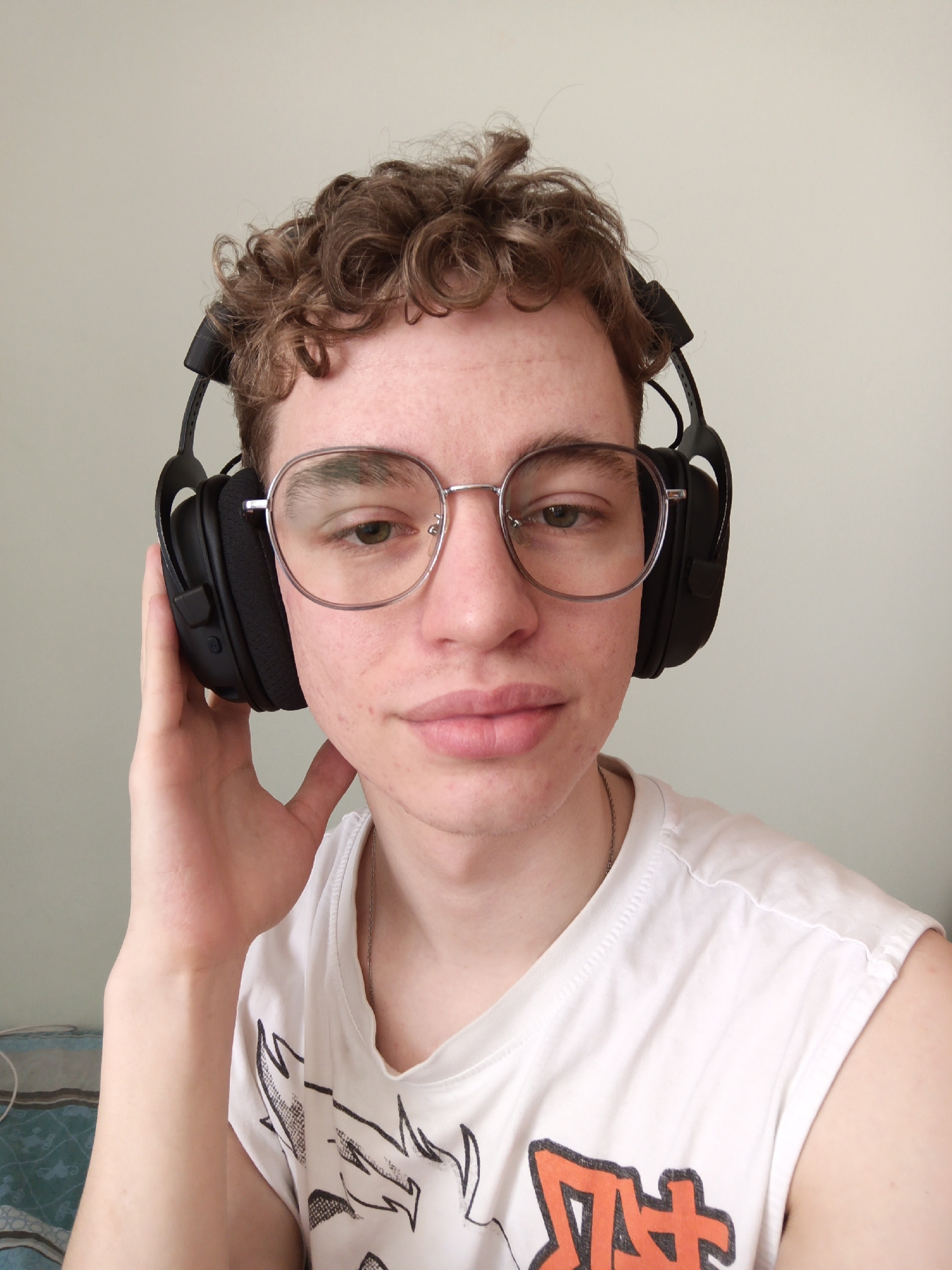 A young man is centered in a portrait-style shot. He has fair skin, curly brown hair, and wears round, silver-rimmed glasses. He is wearing black over-ear headphones and holding one side to his ear with his left hand. He's wearing a white sleeveless shirt with a graphic design on the front. A silver chain necklace is visible around his neck. The background is a plain, light-colored wall. The location, Odintsovo, Russia, is not discernible from this image.