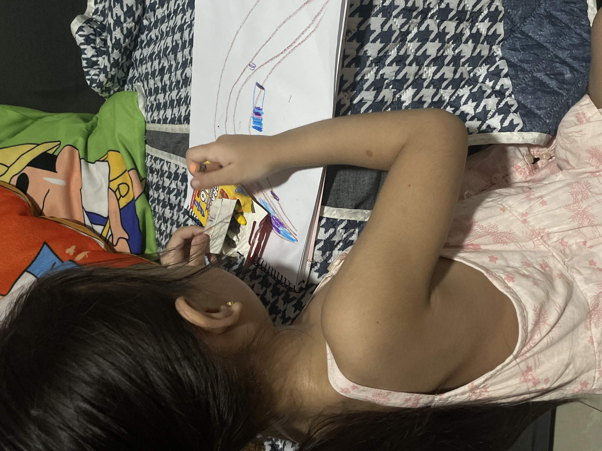 A young girl is shown from the back, sitting and drawing on a large piece of paper. She is wearing a pink dress with a floral pattern. Her hair is dark and pulled back into a ponytail. The girl is holding an orange crayon and appears to be drawing lines and shapes on the paper, which is on a sketchbook. Next to her is a packet of crayons. She is sitting on a bed with a patterned blanket and a pillow with a cartoon character on it. The setting is likely a bedroom in a residential area of Davao City, Philippines.